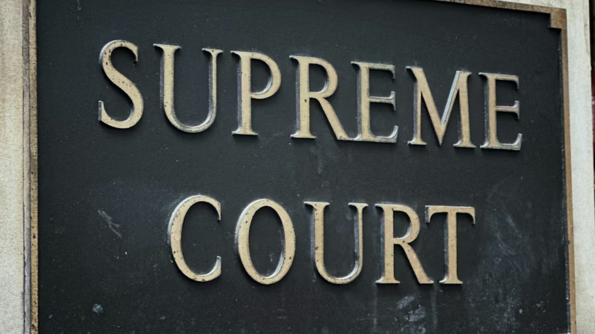 A close-up shot of a dark metallic plaque with the words "SUPREME COURT" embossed in raised, silver capital lettering.