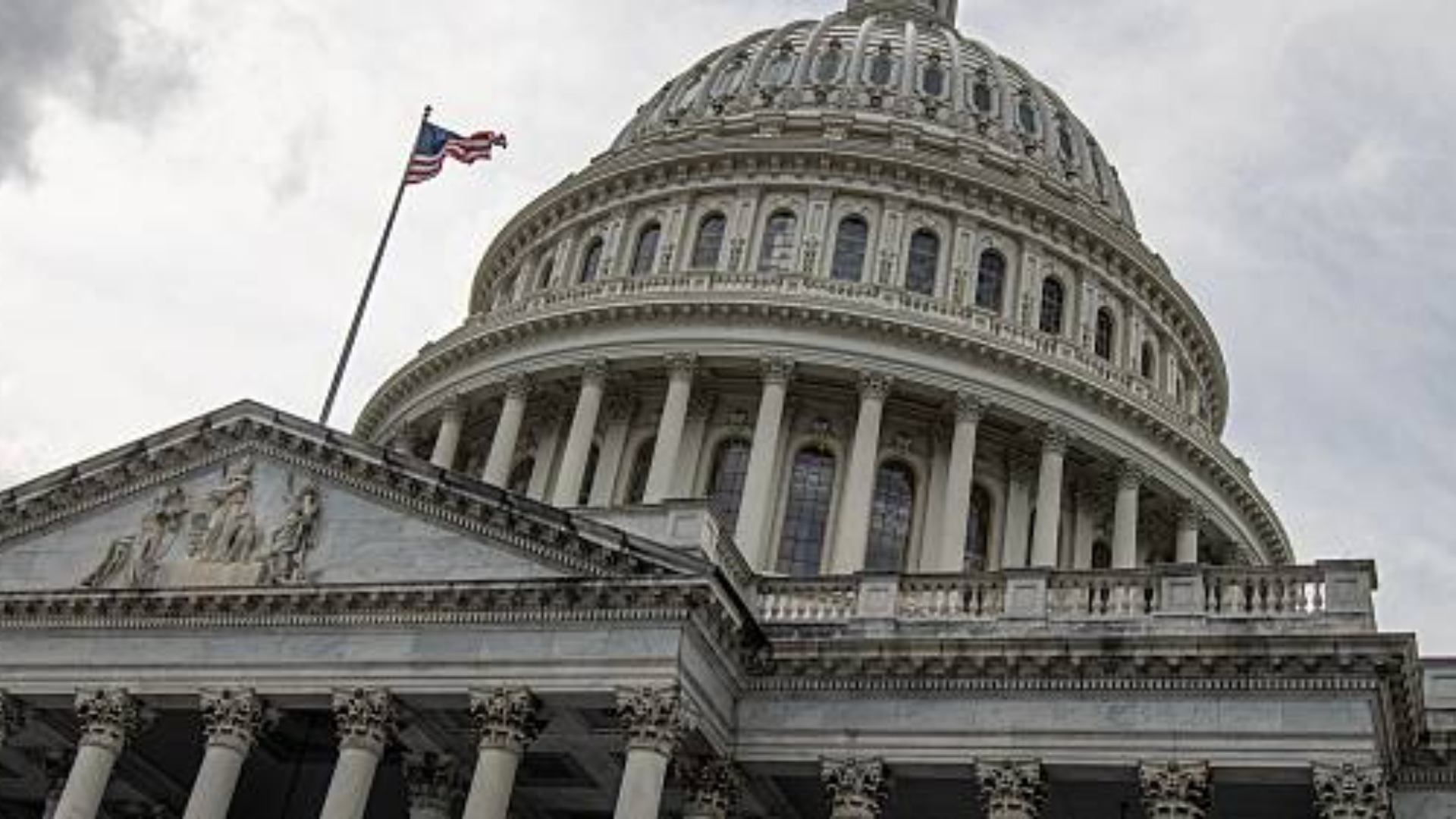 A low-angle exterior view of the United States Capitol building dome in Washington, D.C., with an American flag flying on a pole under a cloudy sky.