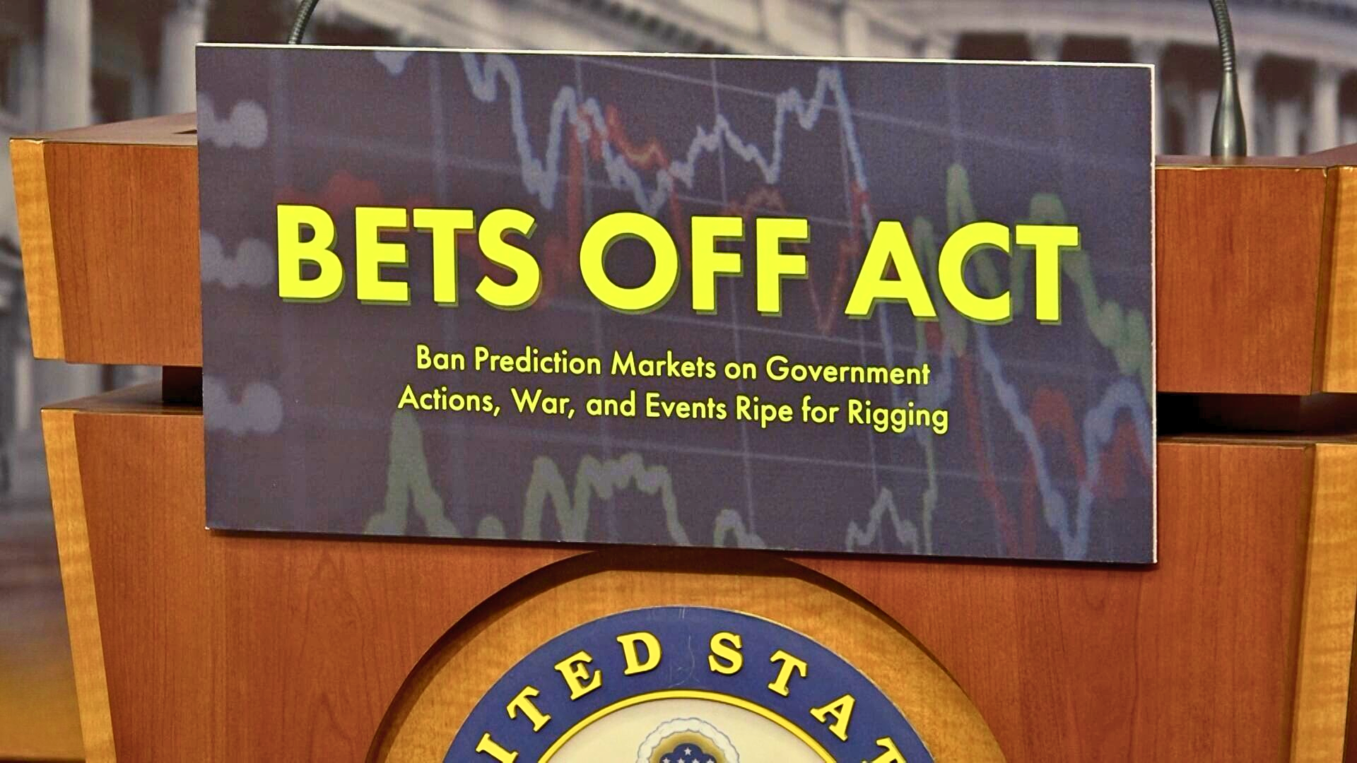 A close-up shot of a sign mounted on a wooden podium that reads "BETS OFF ACT: Ban Prediction Markets on Government Actions, War, and Events Ripe for Rigging."
