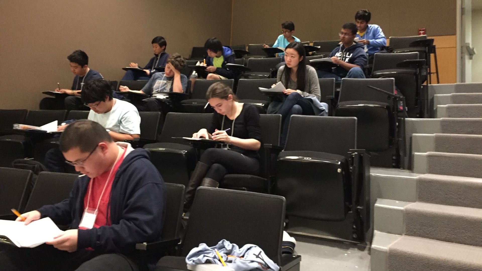 Students seated in a lecture hall write on papers during what appears to be a supervised exam or academic competition.