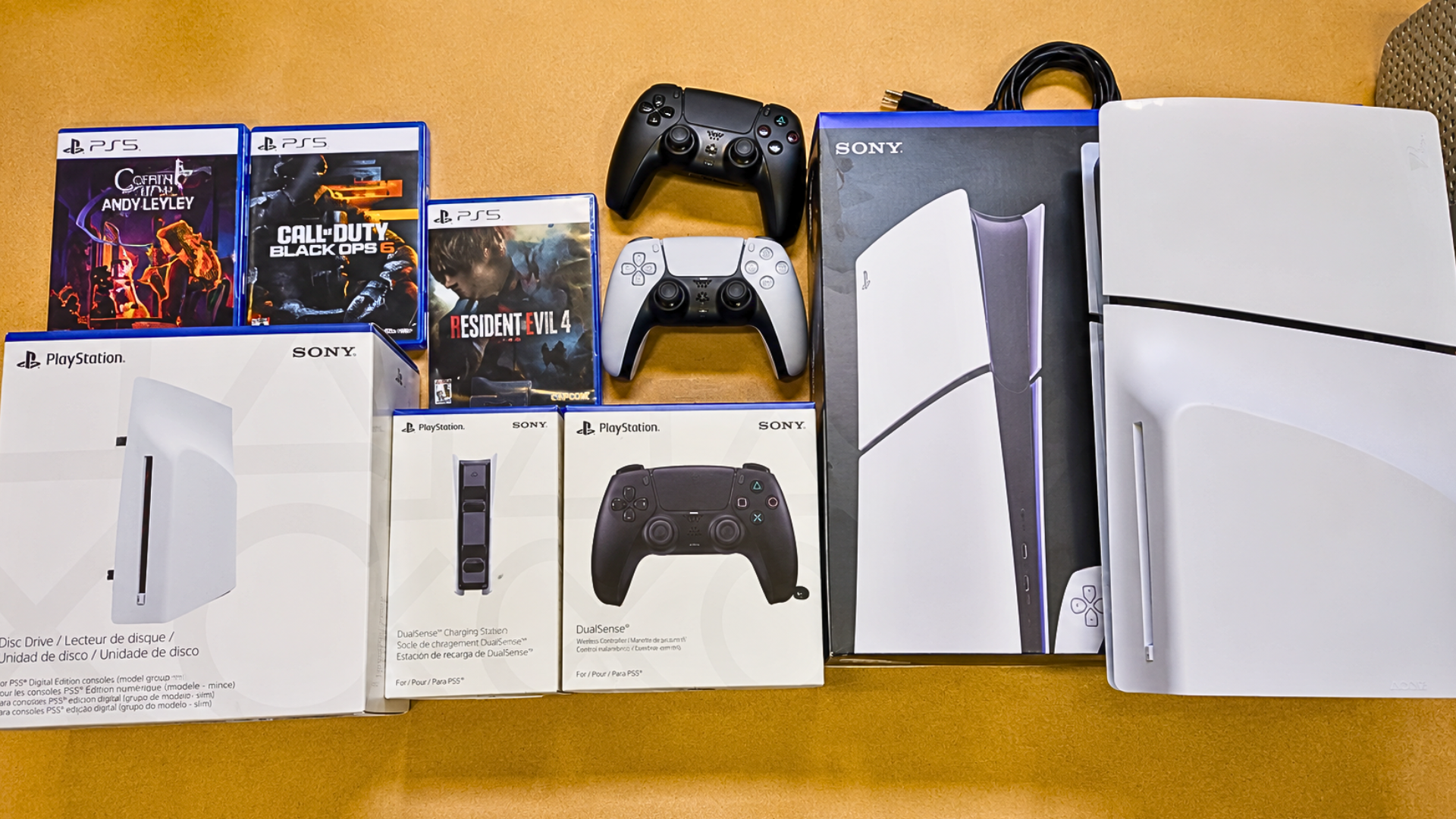 PlayStation 5 console and accessories displayed on a table with PS5 game cases, DualSense controllers, charging dock, and unopened accessory boxes.