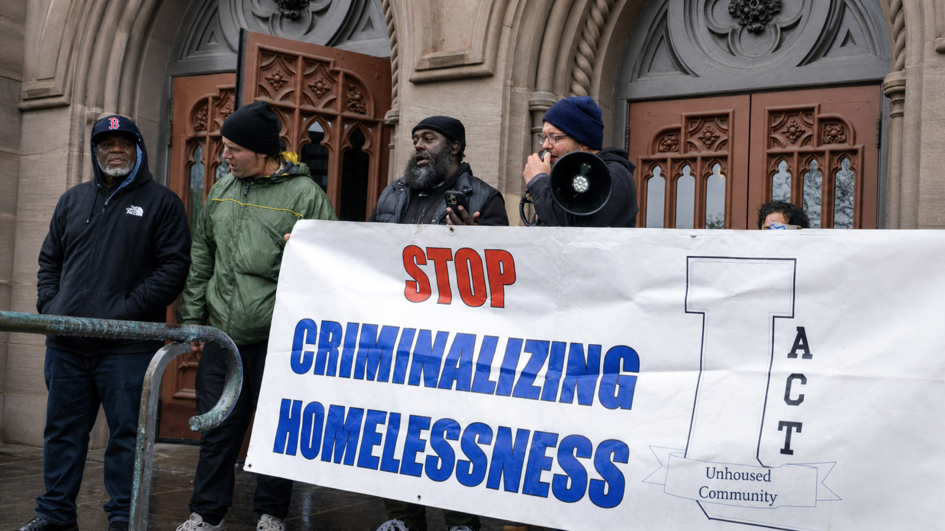 Protesters holding banner reading “Stop criminalizing homelessness” outside a building entrance.