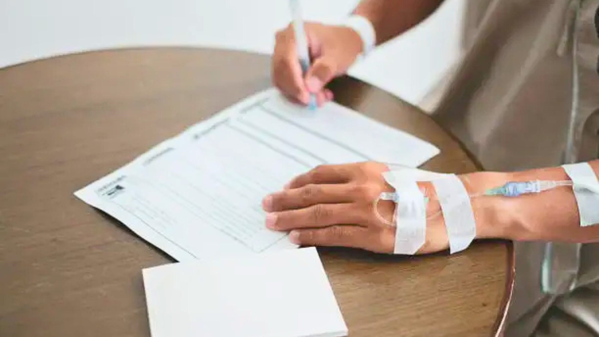 Hospital patient with an IV line signing medical paperwork at a table.
