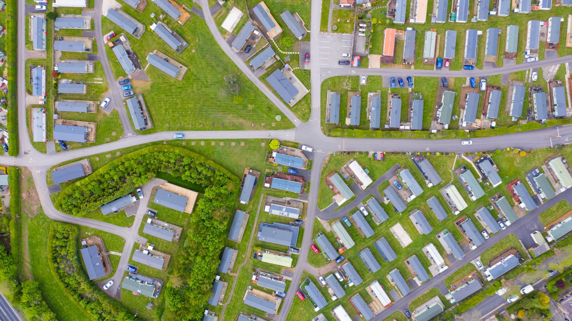 erial view of a large mobile home park with rows of rectangular homes arranged along curved roads and green lawns. Cars are parked beside the homes, and clusters of trees and landscaped areas break up the neighborhood layout.