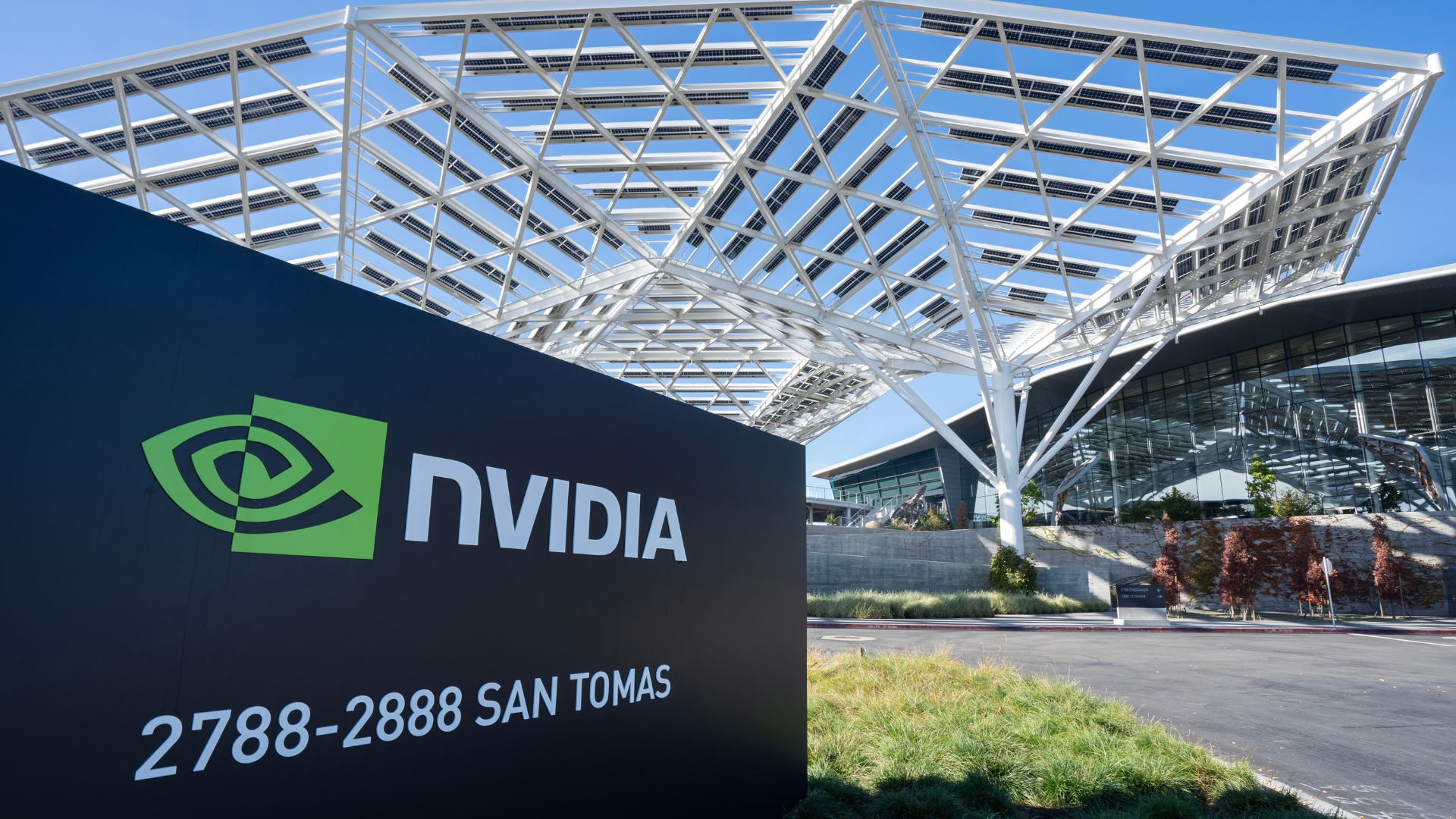 NVIDIA headquarters entrance with a large black sign displaying the NVIDIA logo and the text “2788-2888 SAN TOMAS” in front of a modern glass office building. White steel framework with solar panels stretches overhead against a clear blue sky.