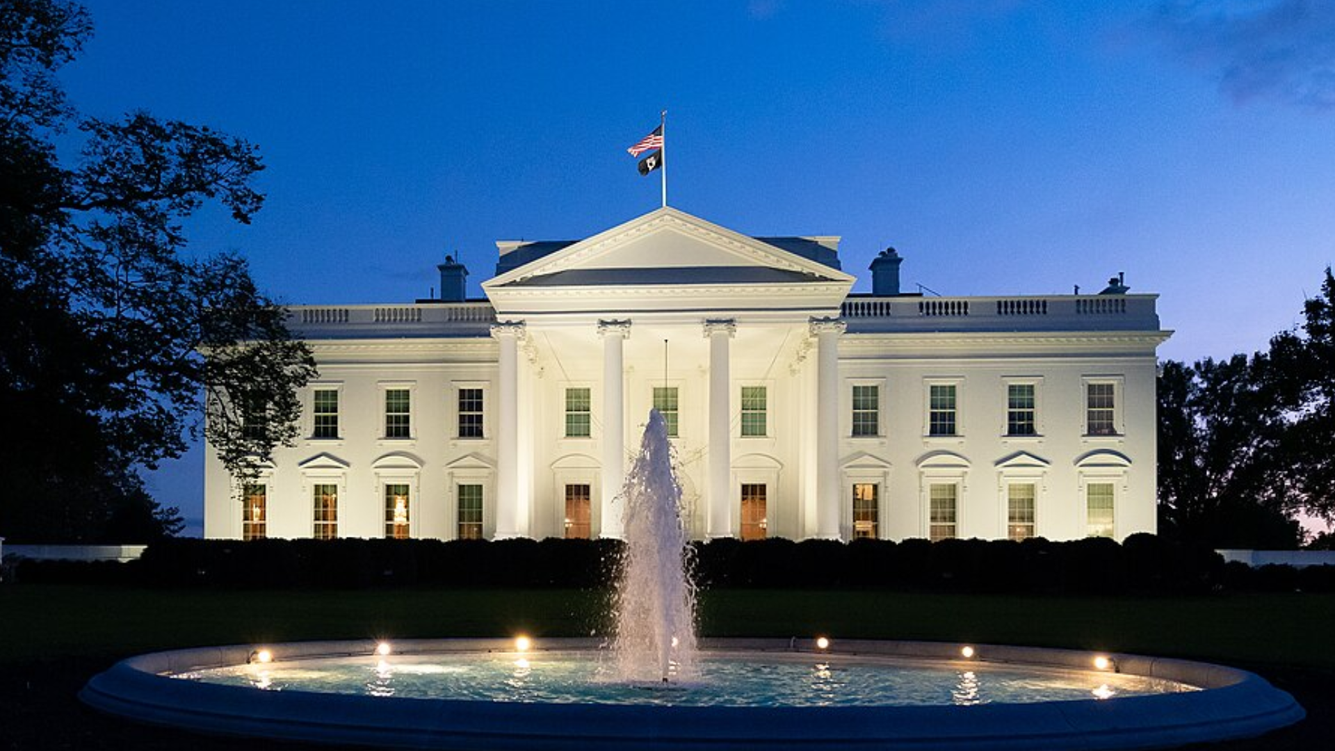 The exterior of the White House brightly illuminated at dusk with a water fountain in the foreground.