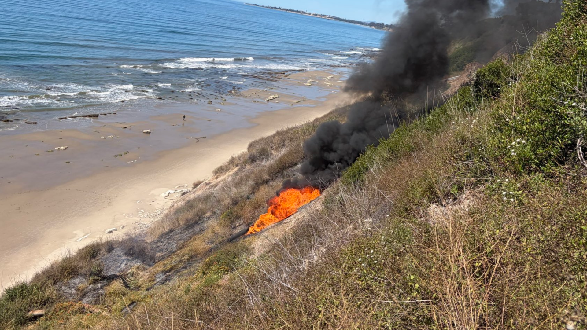 A brush fire burns on a steep coastal cliff next to a sandy beach, sending thick dark smoke into the air.
