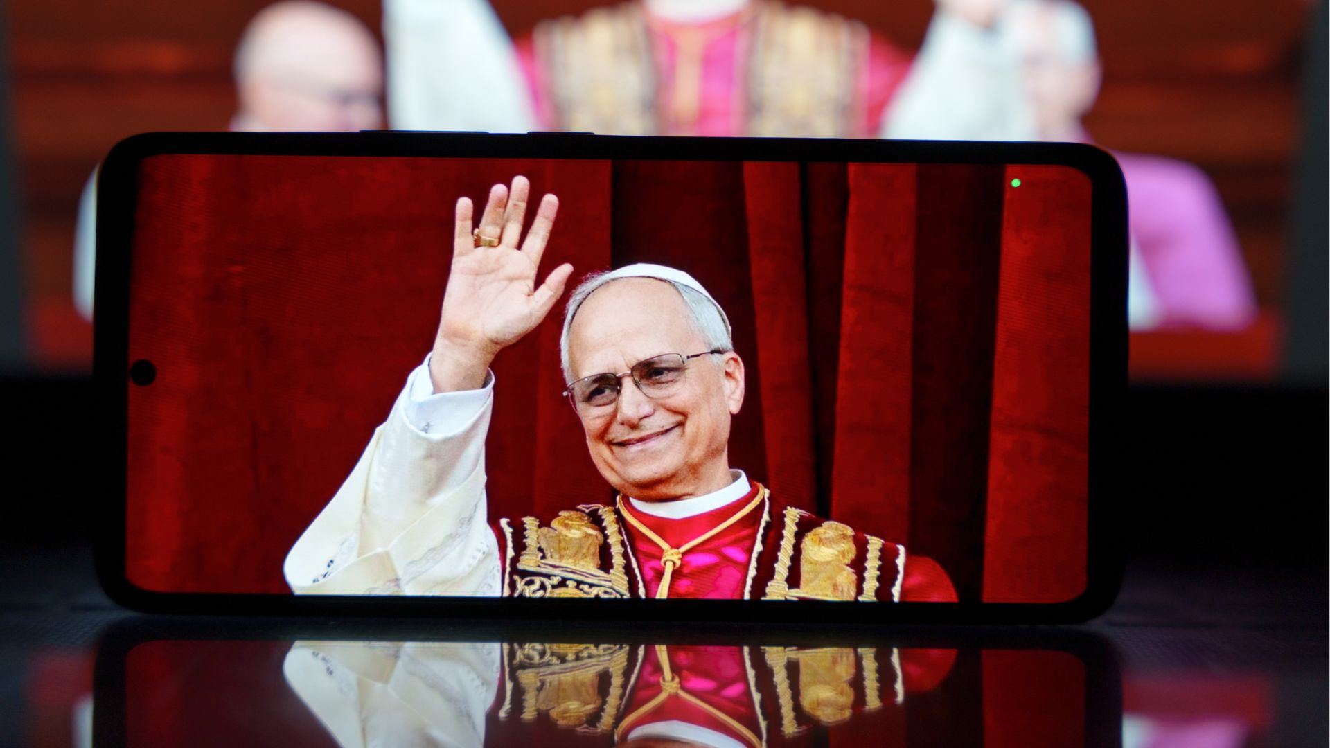 Smartphone displaying Pope Leo XIV waving while wearing red and gold papal vestments against a red curtain backdrop.