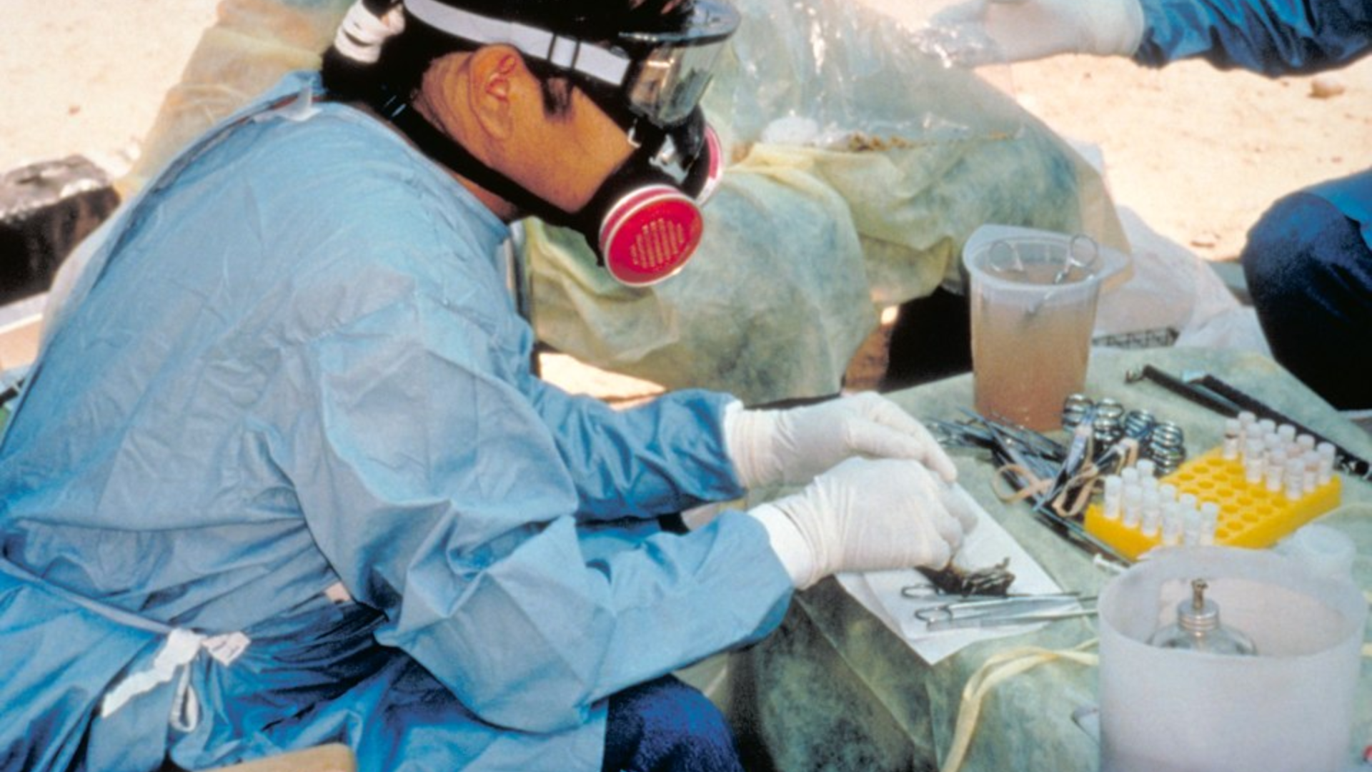 A scientist in full protective gear, including a respirator and goggles, performing an autopsy or sampling in a field laboratory.