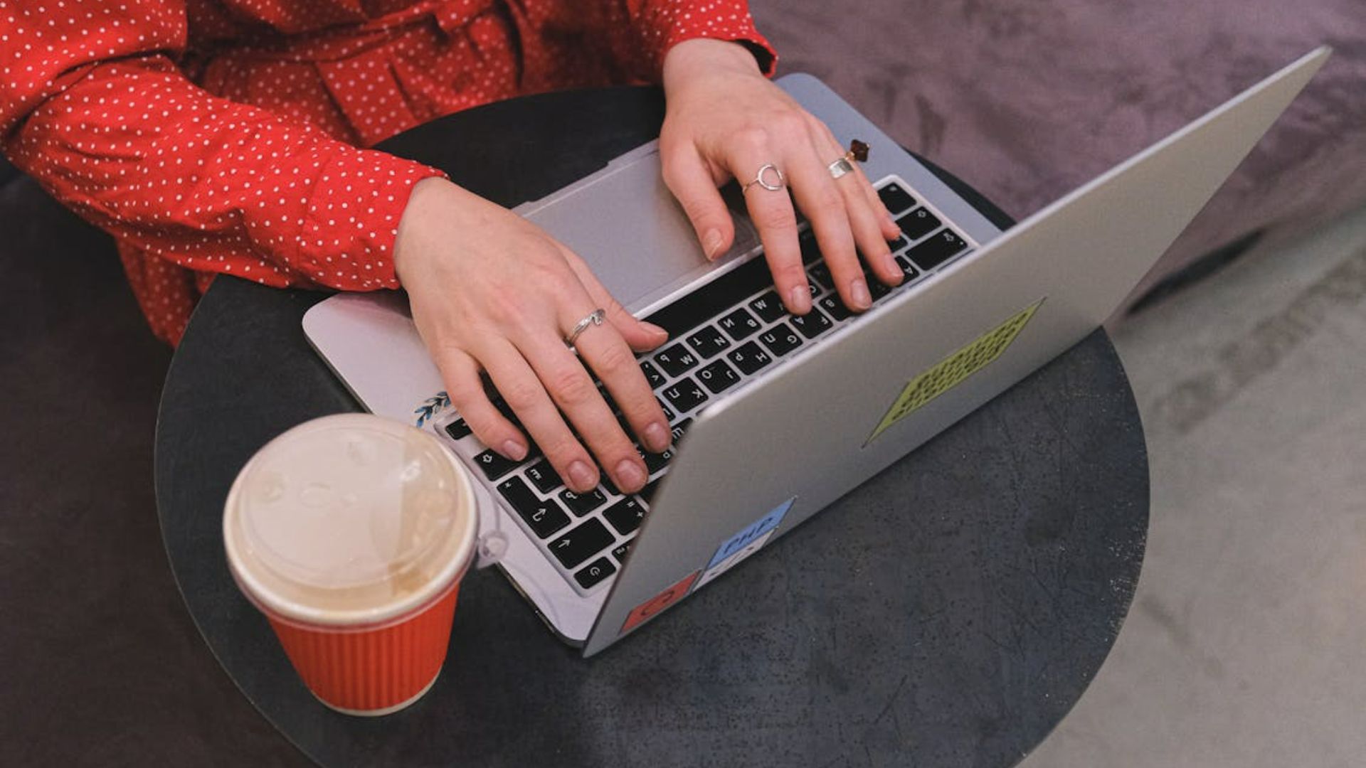 A woman in a red polka dot dress typing on a laptop with coffee nearby.