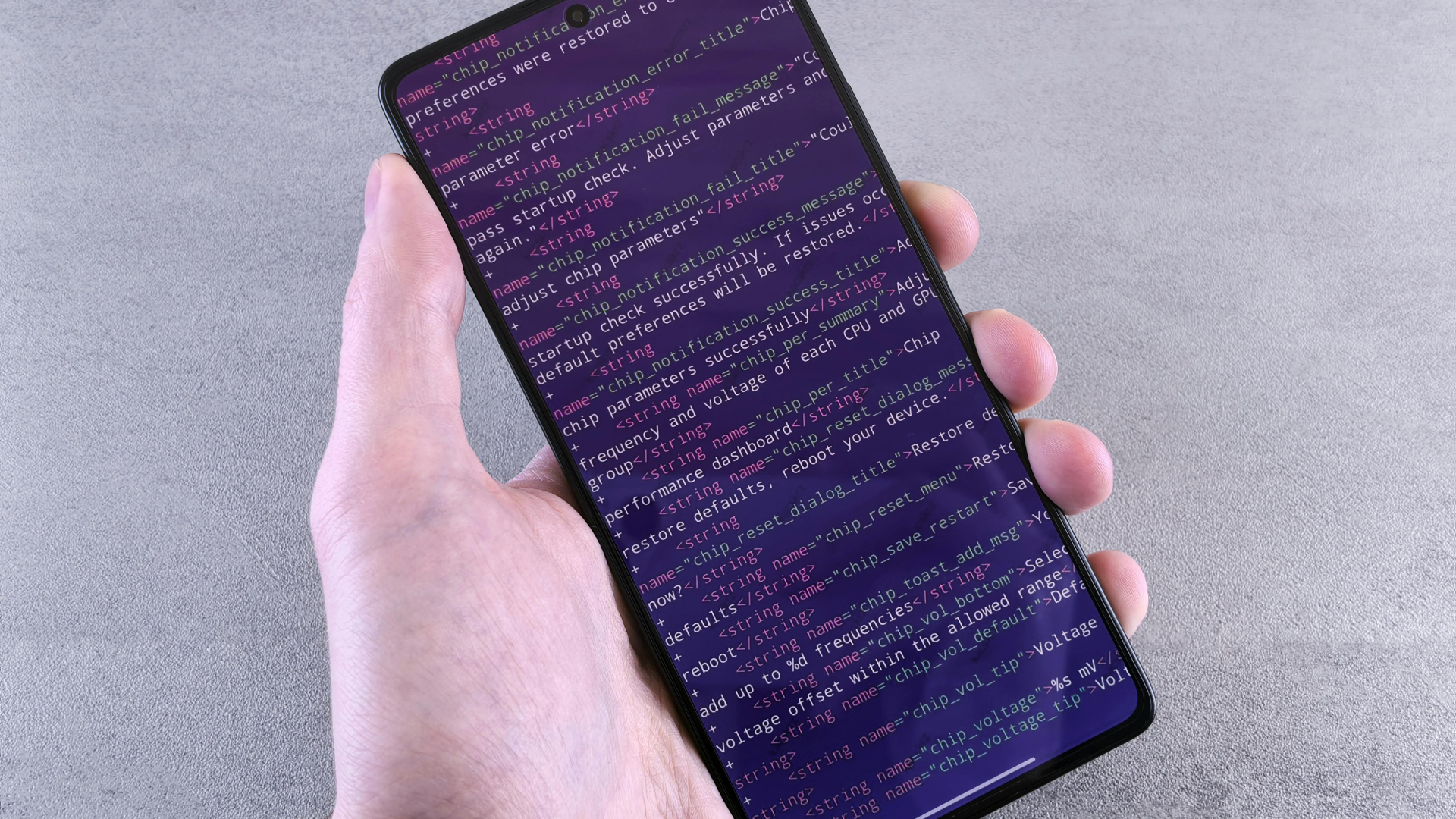 A person's hand holds a black smartphone displaying rows of pink, white, and purple XML code on the screen against a textured grey background.