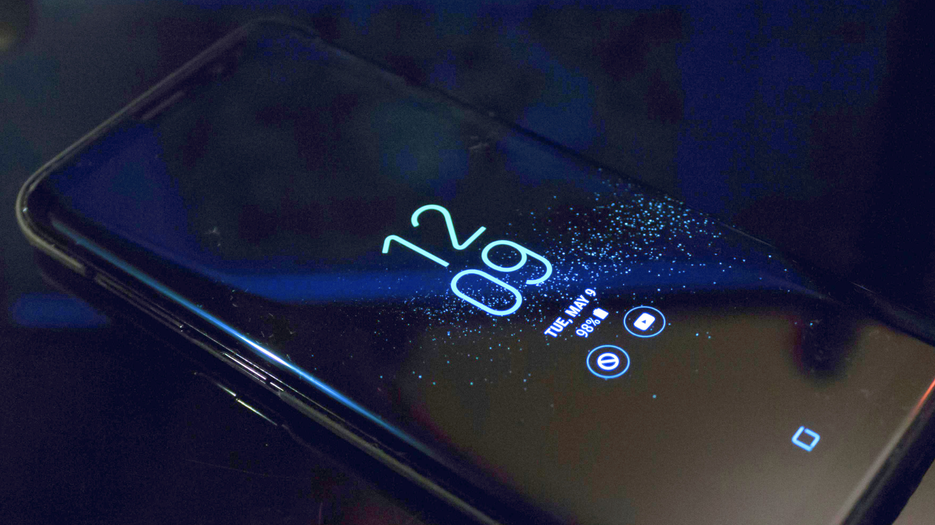 A smartphone lies on a dark surface in a dimly lit room, with its always-on display showing a teal digital clock reading 12_09 alongside minor status icons.