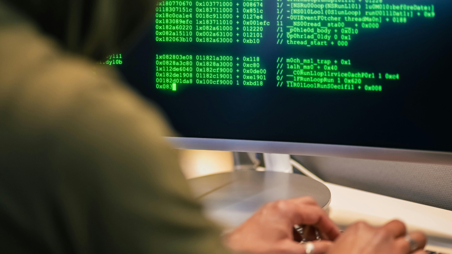 An over-the-shoulder view of a person wearing a green hoodie typing on a keyboard, with bright green computer code filling the monitor screen in front of them.