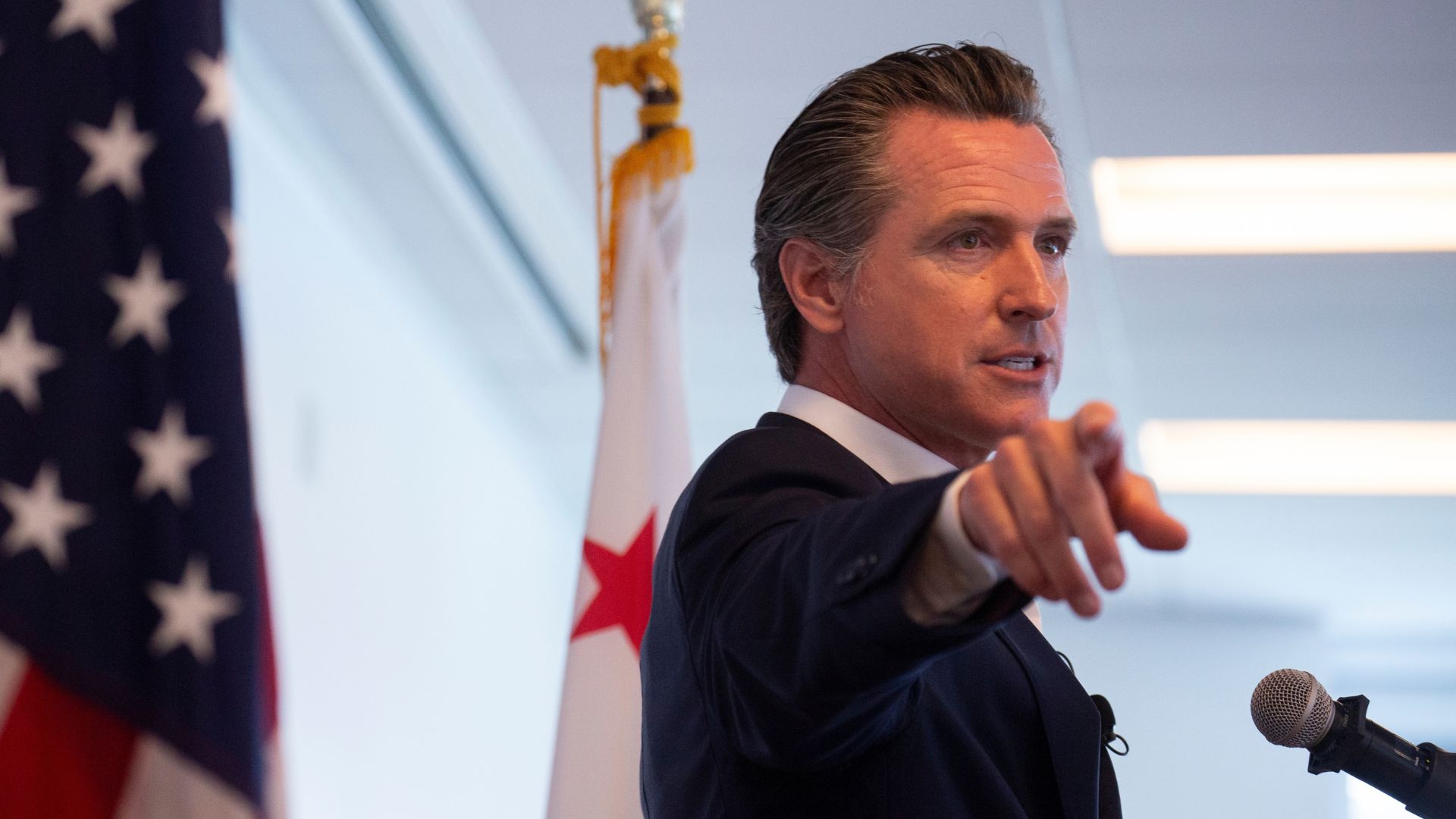 California Governor Gavin Newsom speaks at a podium and points toward the camera, with an American flag and a California state flag visible in the background.