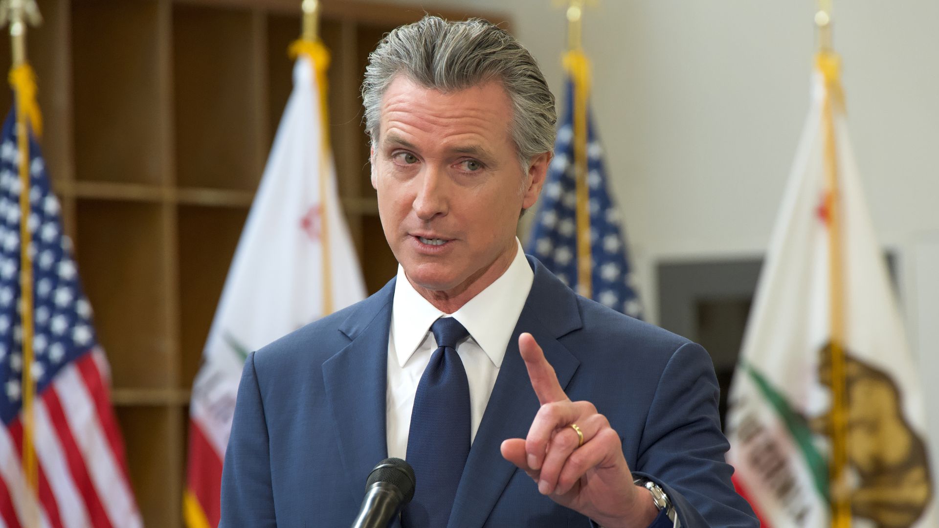 California Governor Gavin Newsom gestures with one finger raised while speaking at a microphone.