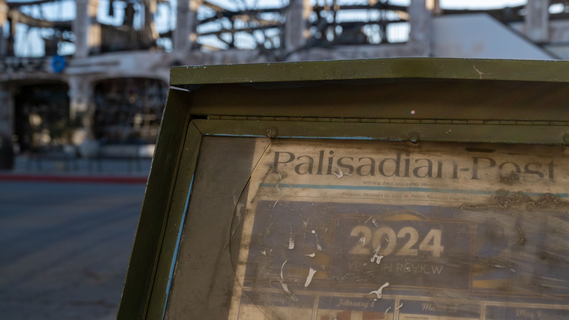 A weathered Palisadian-Post newspaper box stands in front of a fire-gutted building stripped to its bare frame.