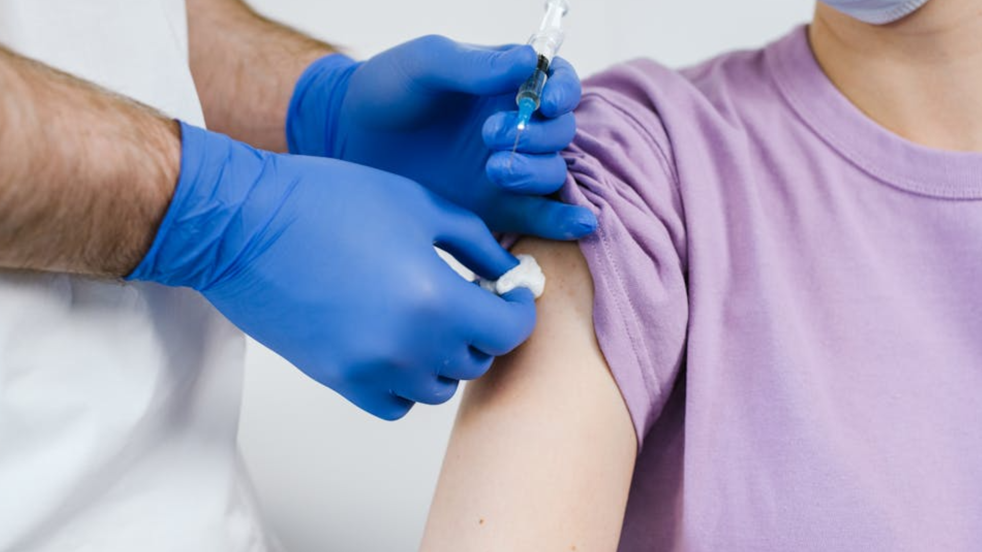 A close-up shot of a healthcare provider in white scrubs and blue nitrile gloves cleaning a patient's upper arm with an alcohol swab before administering an injection with a clear.