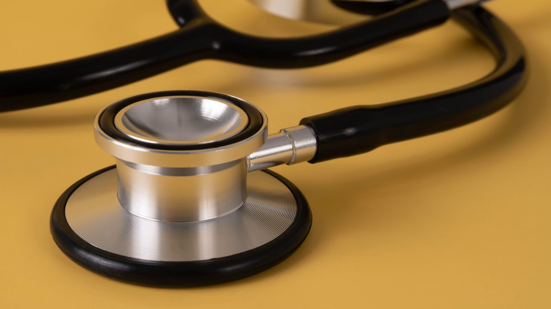A close-up shot of a stainless-steel stethoscope chestpiece with its black rubber non-chill rim, resting on a flat, solid mustard-yellow background with the black binaural tubing.
