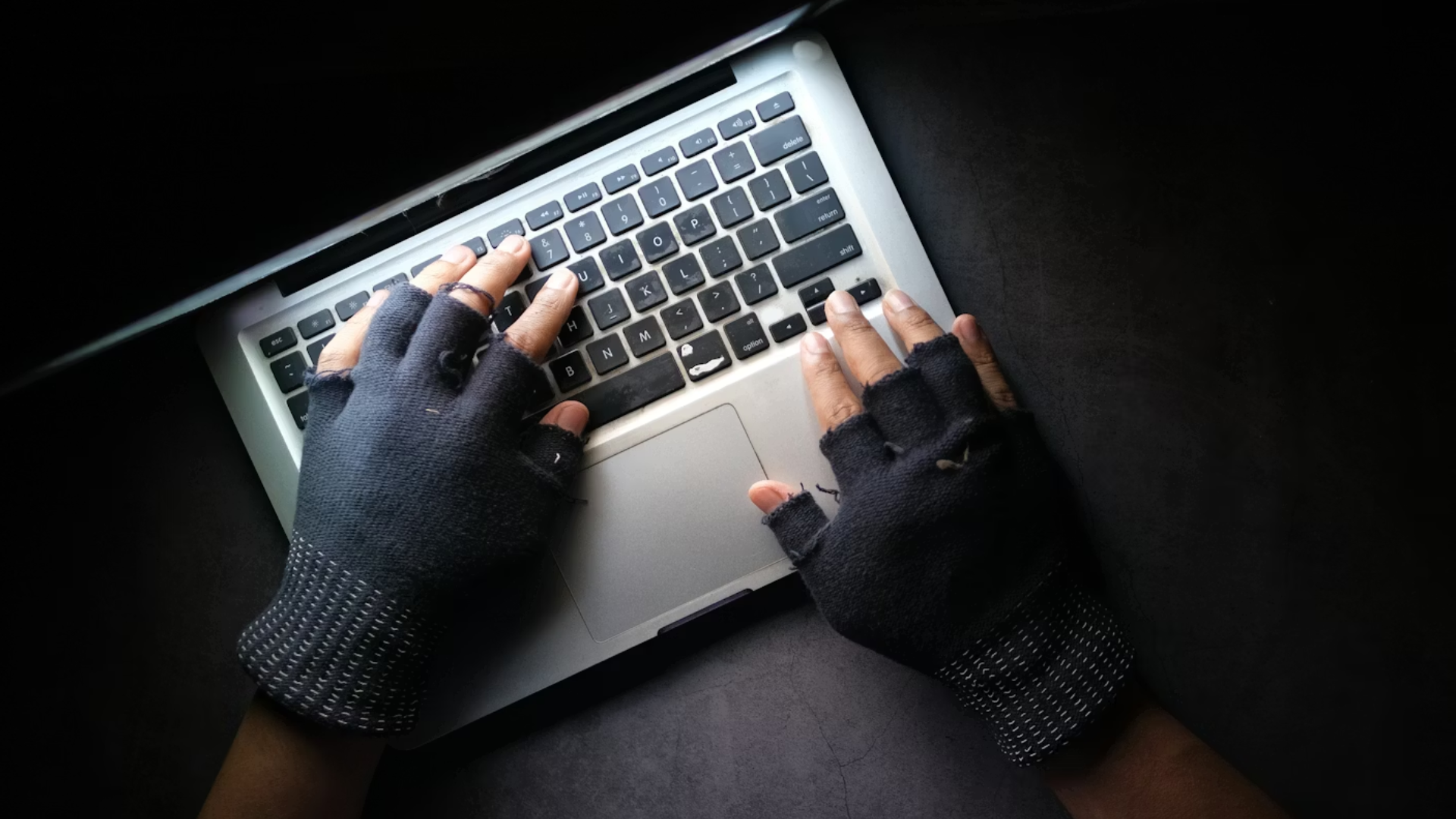 An overhead shot of a person wearing black fingerless gloves typing on a silver laptop in a dark, moody setting.