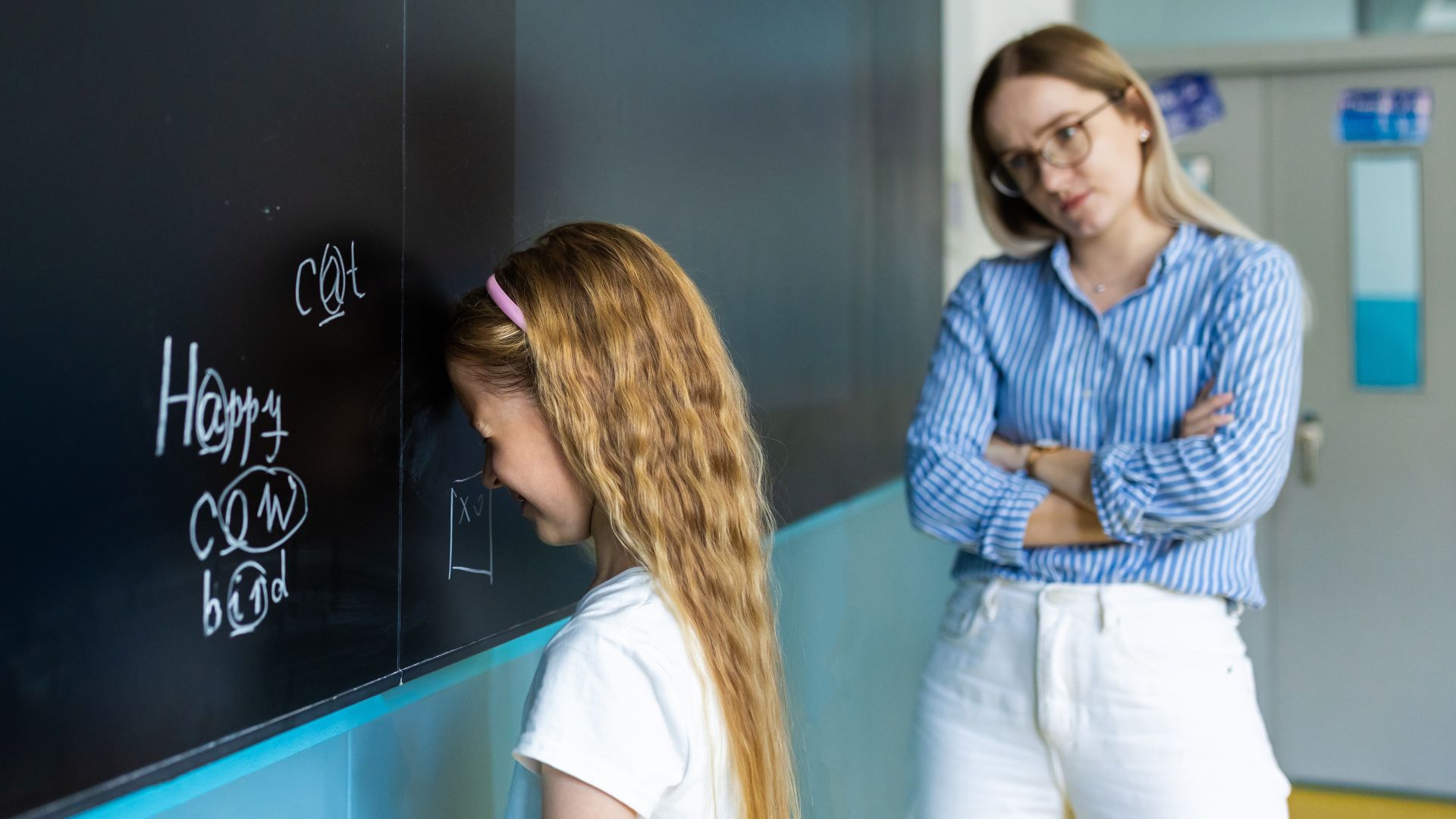 Young girl stands facing a classroom chalkboard with her head lowered while an adult teacher watches from behind with crossed arms.