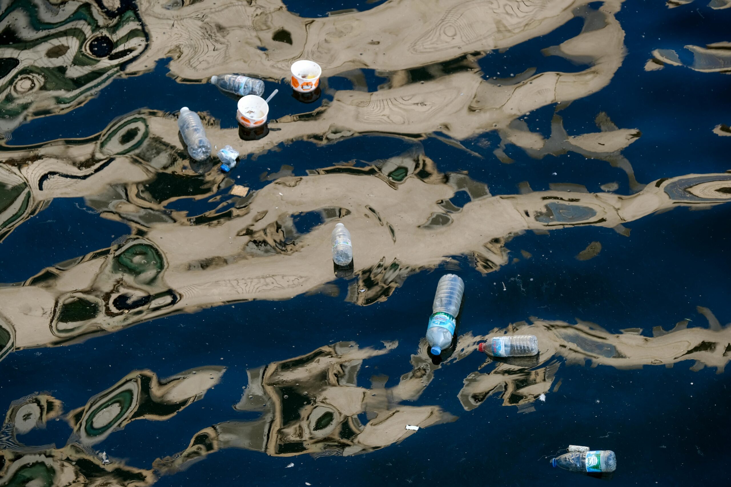 Plastic bottles and trash float across polluted water covered in oily patterns, illustrating environmental contamination and concerns over toxic chemical pollution in waterways.