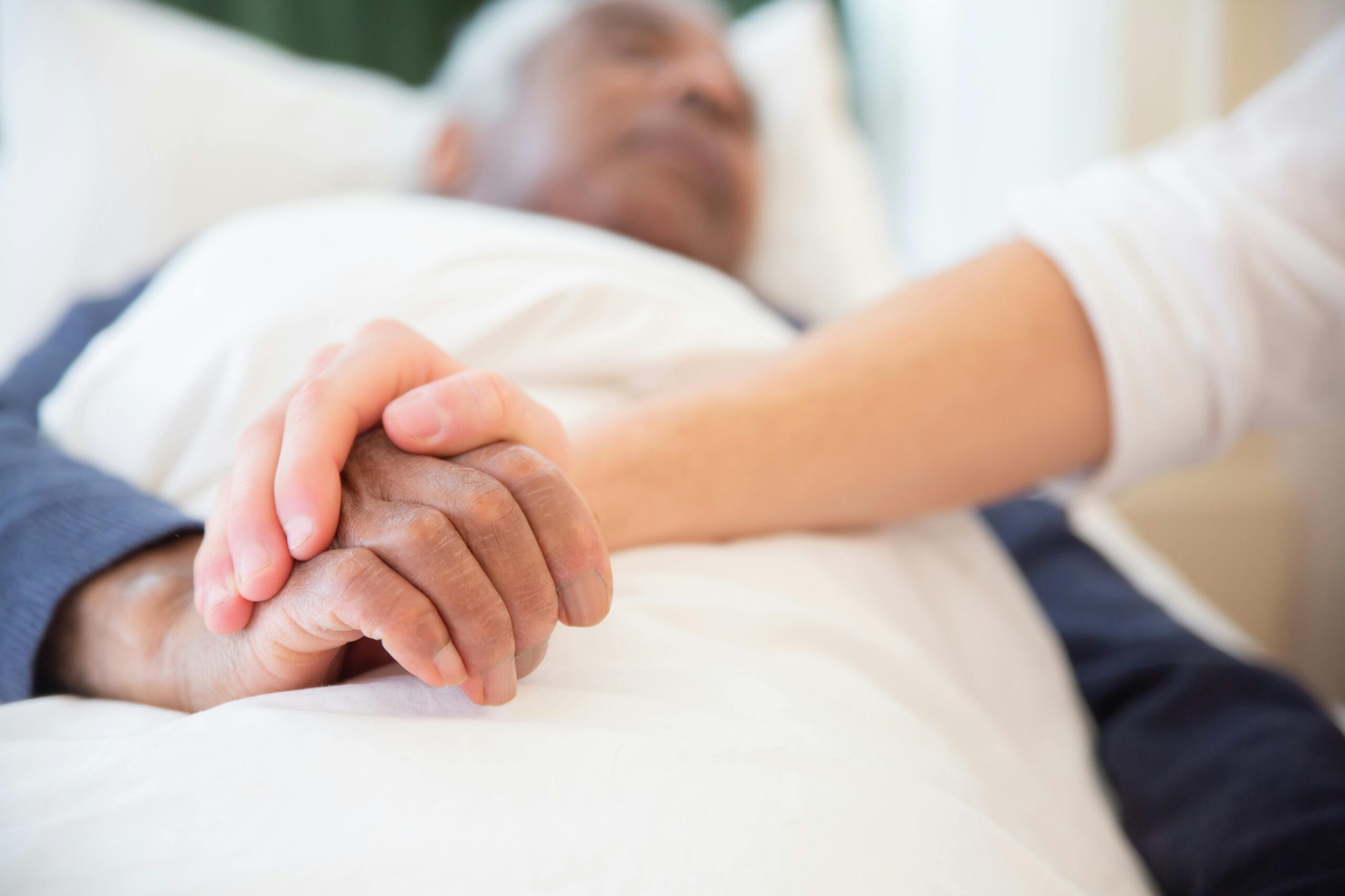 Caregiver holds an elderly patient’s hand while providing bedside hospice or home healthcare support.