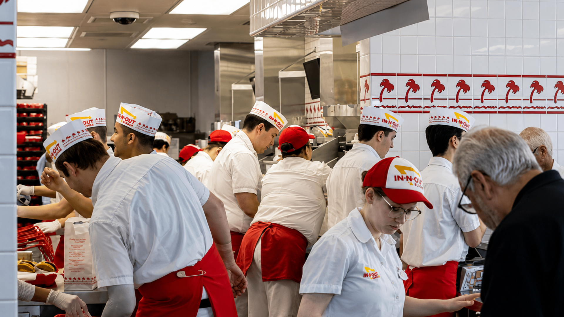 A busy kitchen crew wearing white uniforms, red aprons, and paper hats preparing food inside an In-N-Out Burger restaurant.