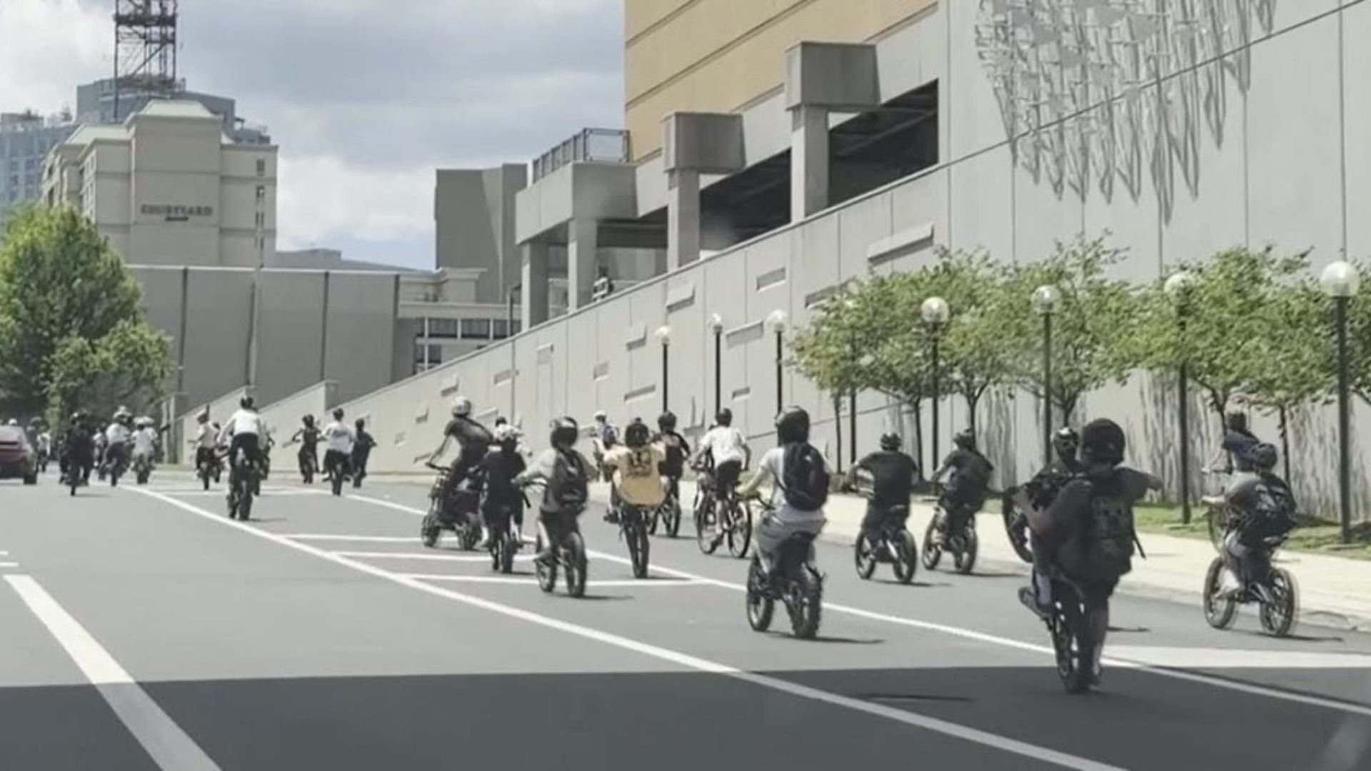 A large group of teenagers ride bicycles and e-bikes together down a city street, filling multiple lanes as they pass office buildings and sidewalks. The scene reflects the large gathering that prompted safety concerns after nearly 100 teens on bikes and e-bikes swarmed a Virginia street.