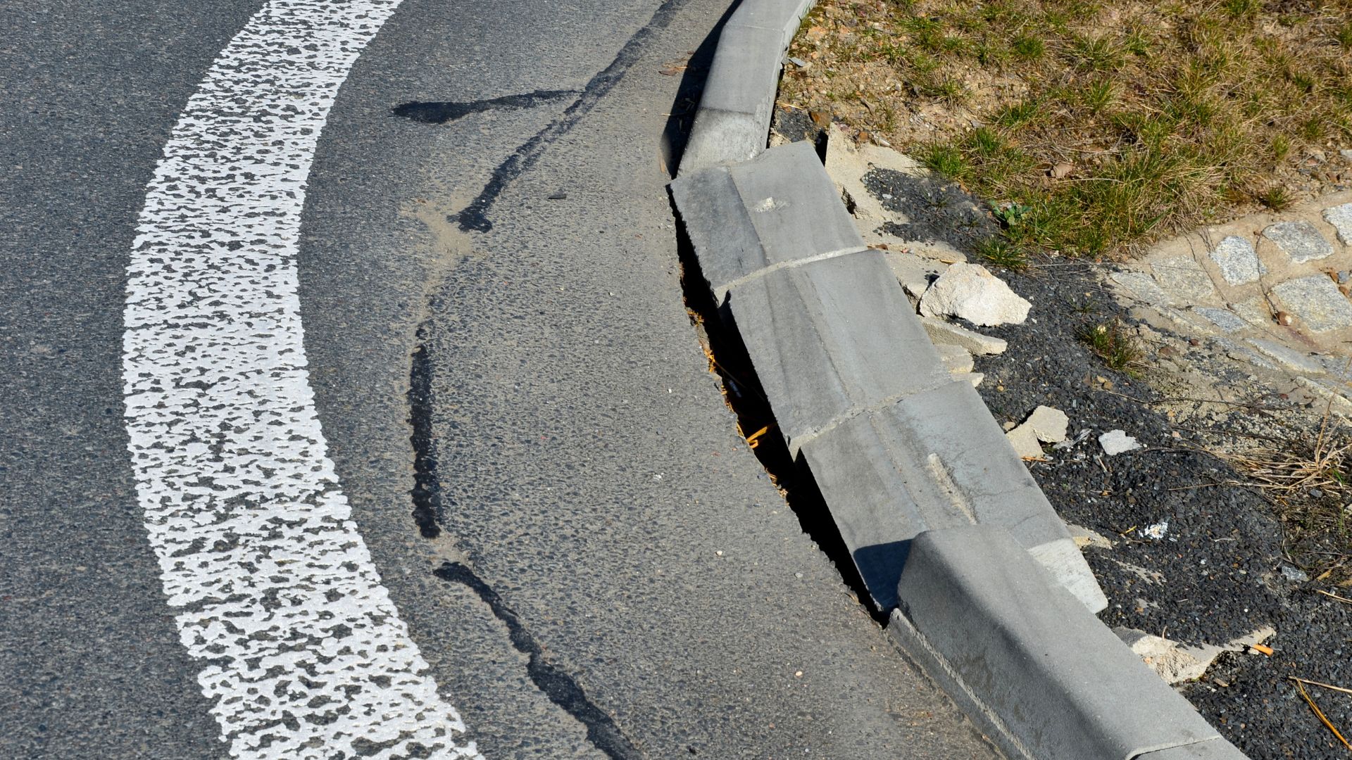 Damaged concrete curb with broken sections pushed out of place beside the edge of a paved roadway. The cracked curb illustrates infrastructure damage that can result from traffic impacts and raises broader questions about street safety.
