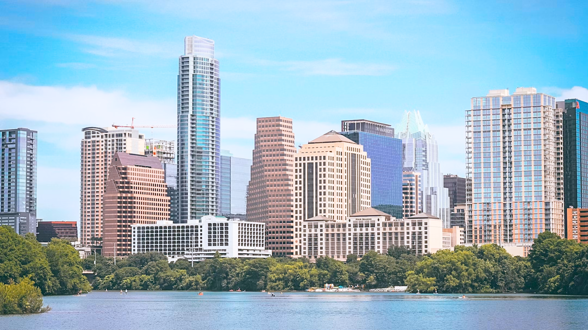 The downtown Austin city skyline rising above a tree-lined river on a clear day.