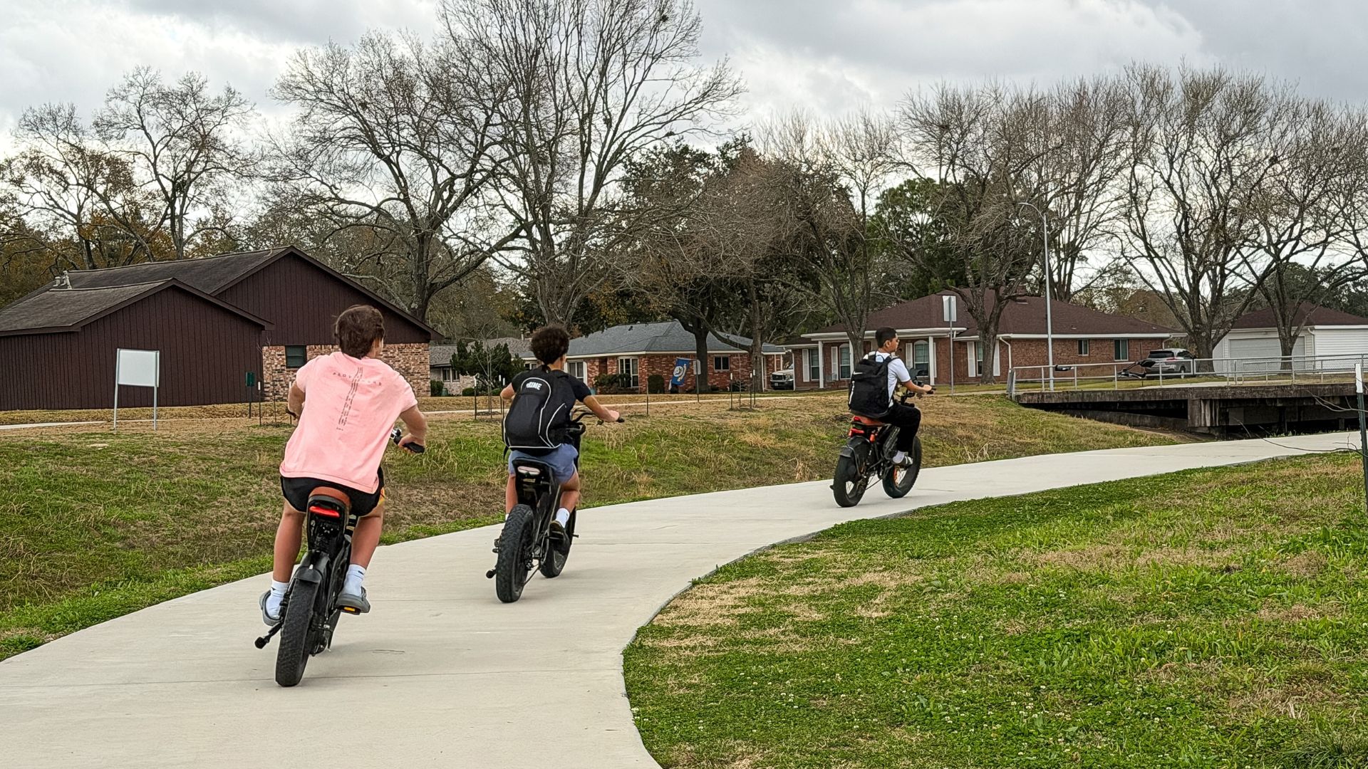 Three teenagers ride e-bikes along a shared-use concrete trail through a suburban neighborhood bordered by grass, homes, and leafless trees.