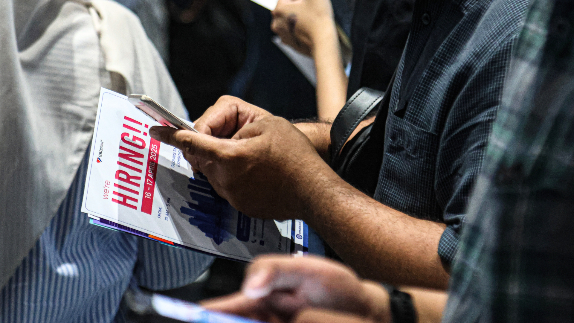 A close-up of a person holding a smartphone and a "we're HIRING!!" flyer while standing in a crowd.