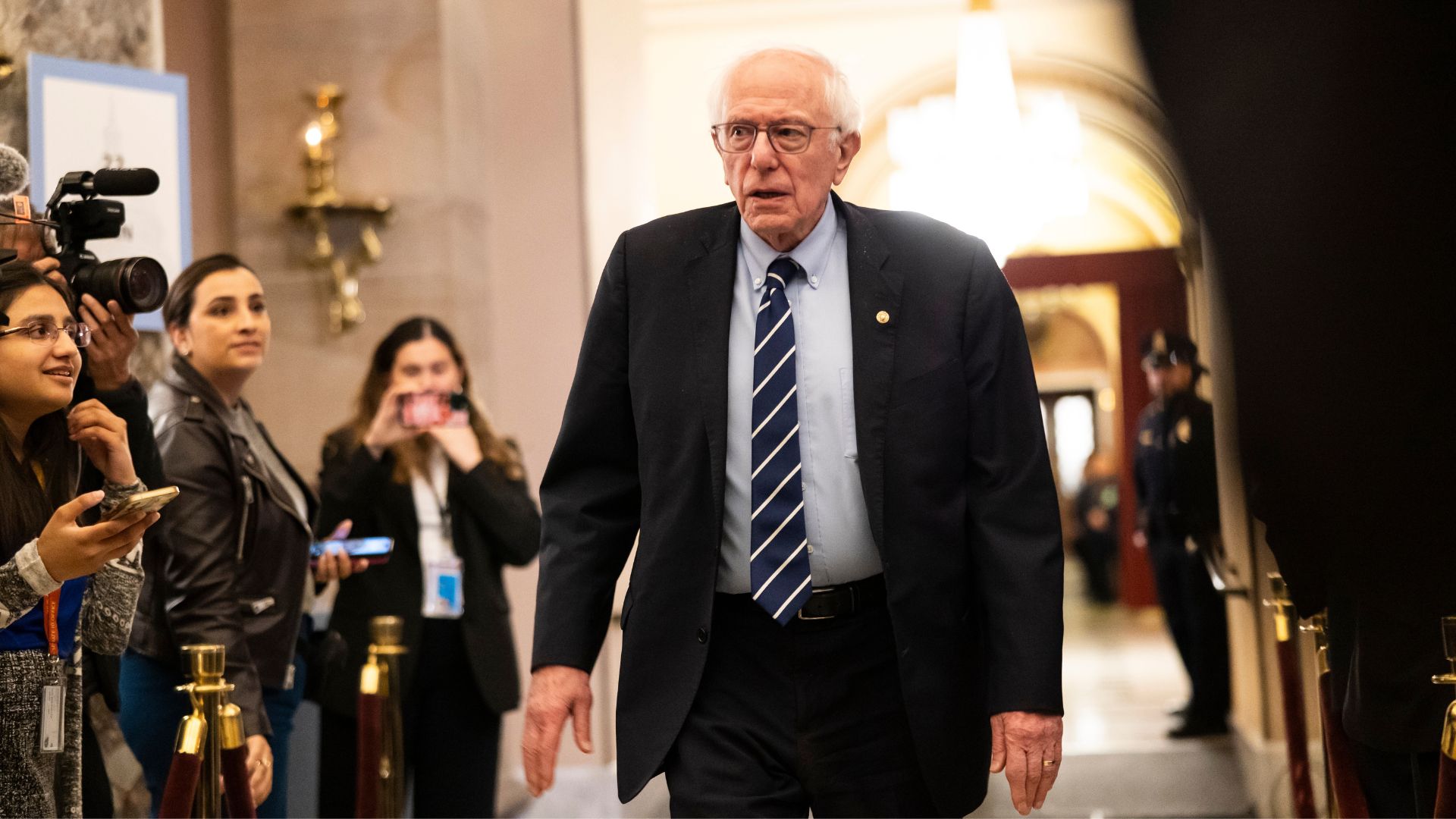 Bernie Sanders walks through a government building hallway as reporters and photographers gather around him.