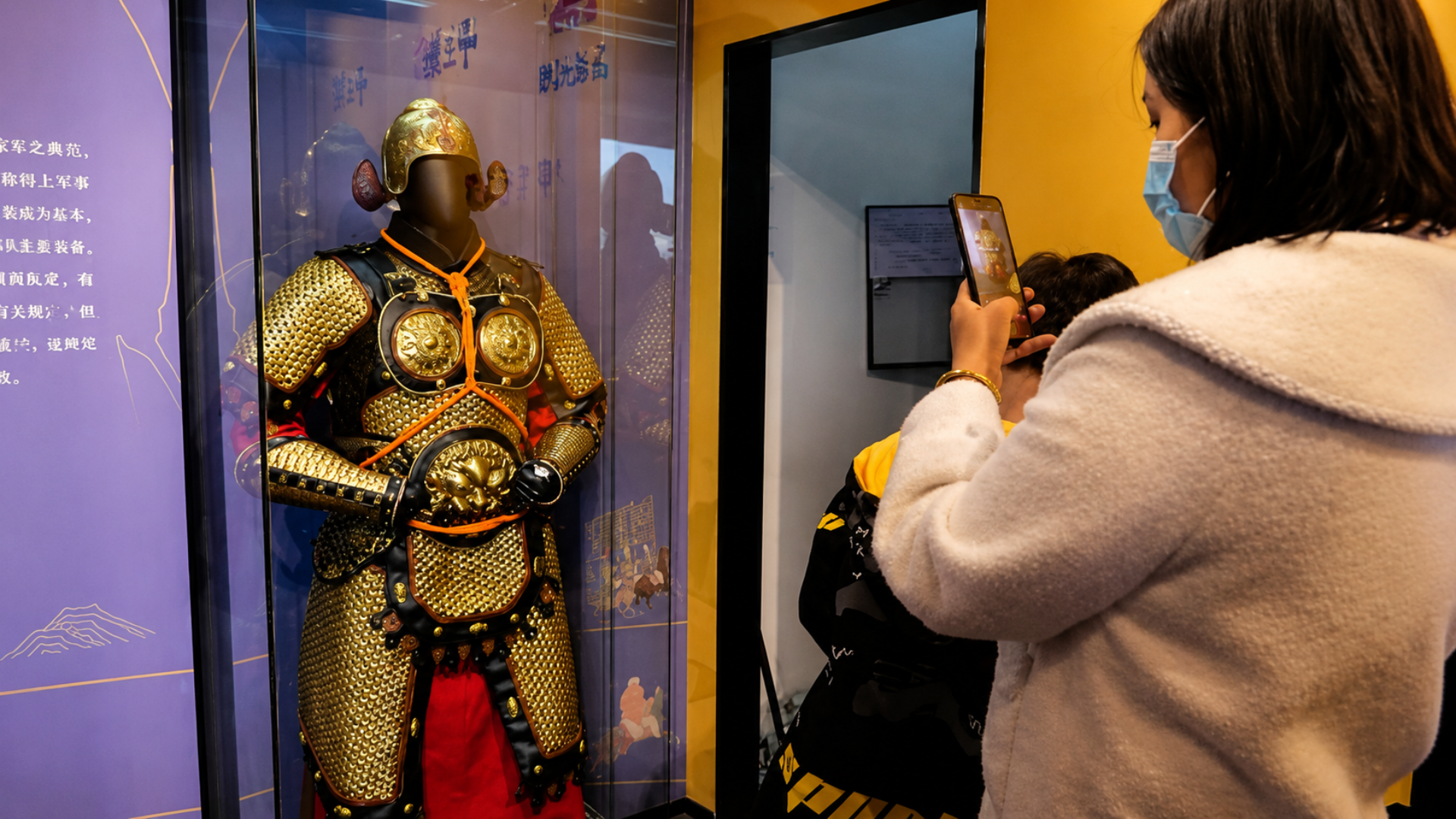 Museum visitor photographing a gold armor exhibit with a smartphone.