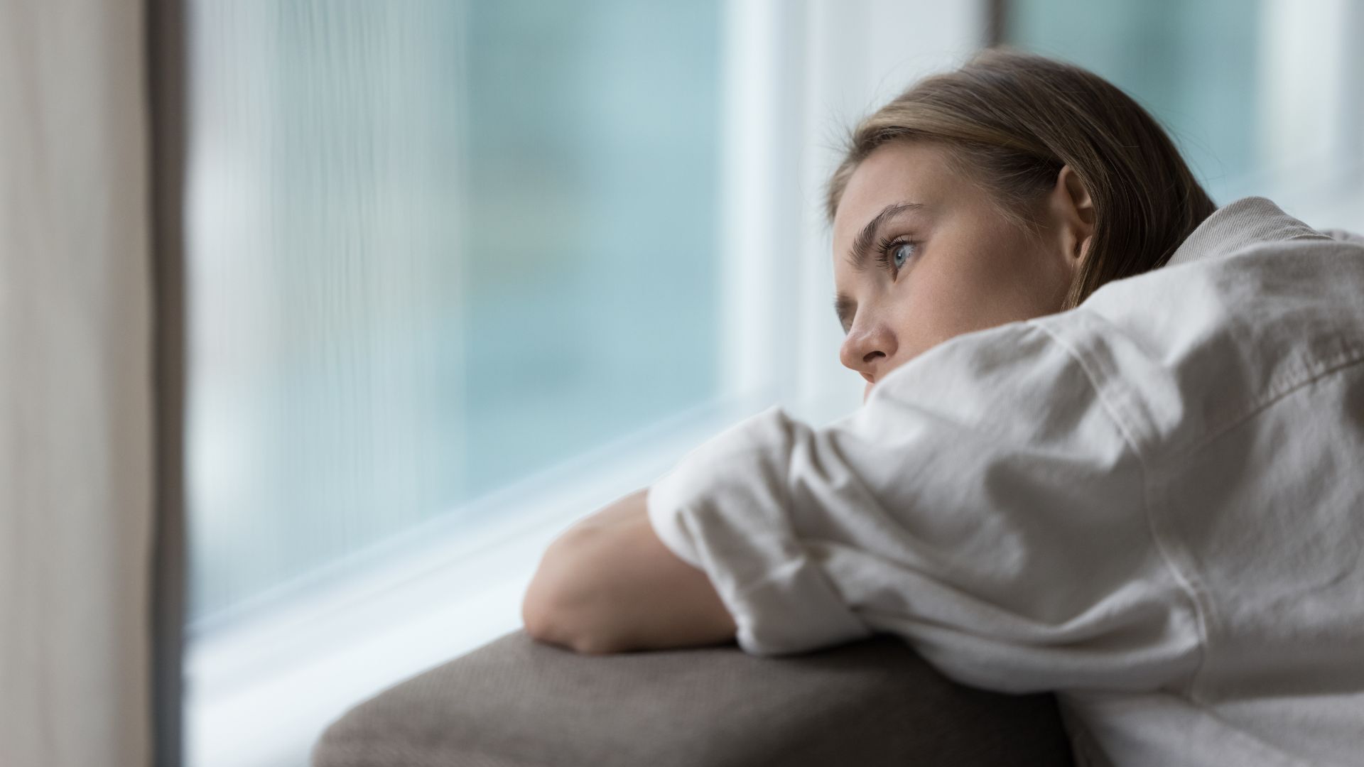 A person looking out through a window with a miserable and contemplative expression, conveying feelings of frustration, sadness, and economic uncertainty.