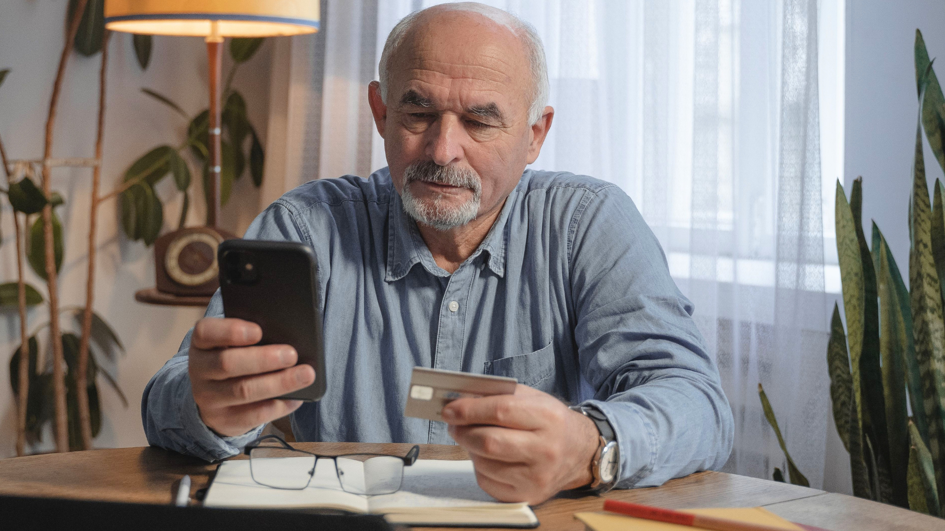 An elderly man with a grey beard sits at a wooden table, holding a black smartphone in his right hand and a silver credit card in his left hand.