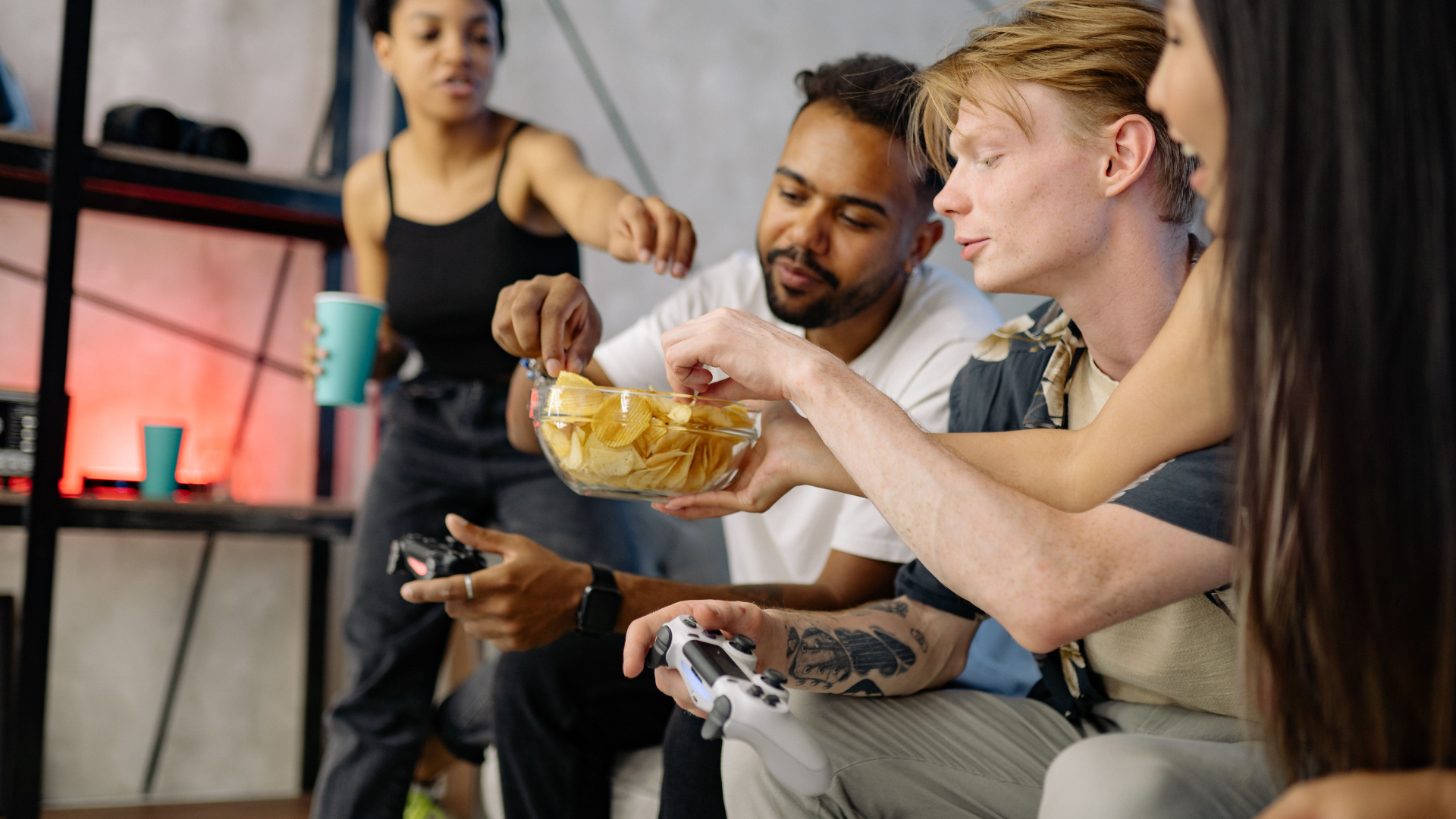 A group of friends sits on a sofa, eating potato chips out of a glass bowl while playing video games with controllers.