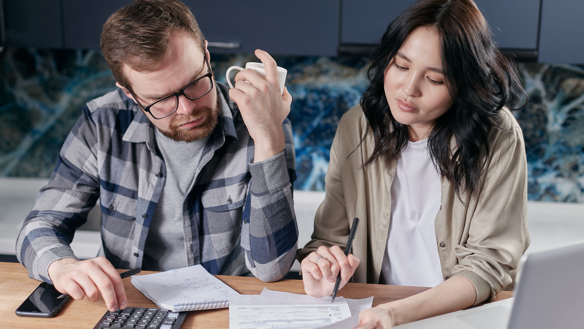 A man and a woman sit at a wooden table looking stressed as they review financial documents alongside a calculator, a laptop, and a notepad.