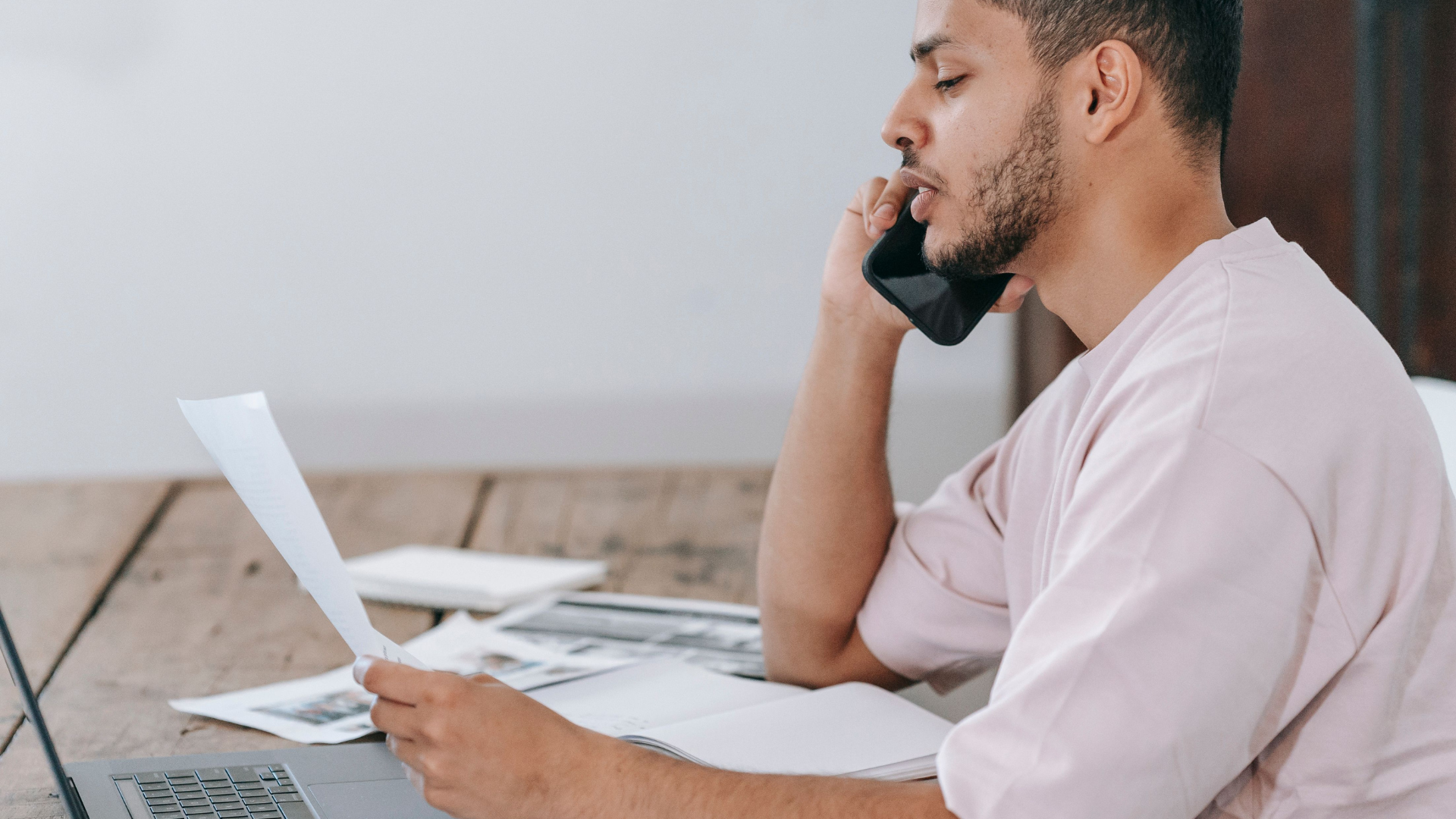 A man sits at a wooden table in front of a laptop, holding a document while speaking on his smartphone.