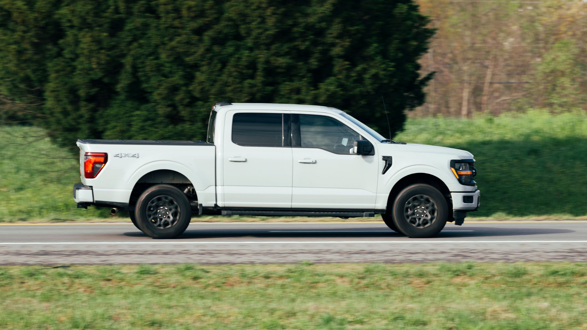 A side view of a white Ford F-150 pickup truck driving on an asphalt road past a large green tree and a grassy field.