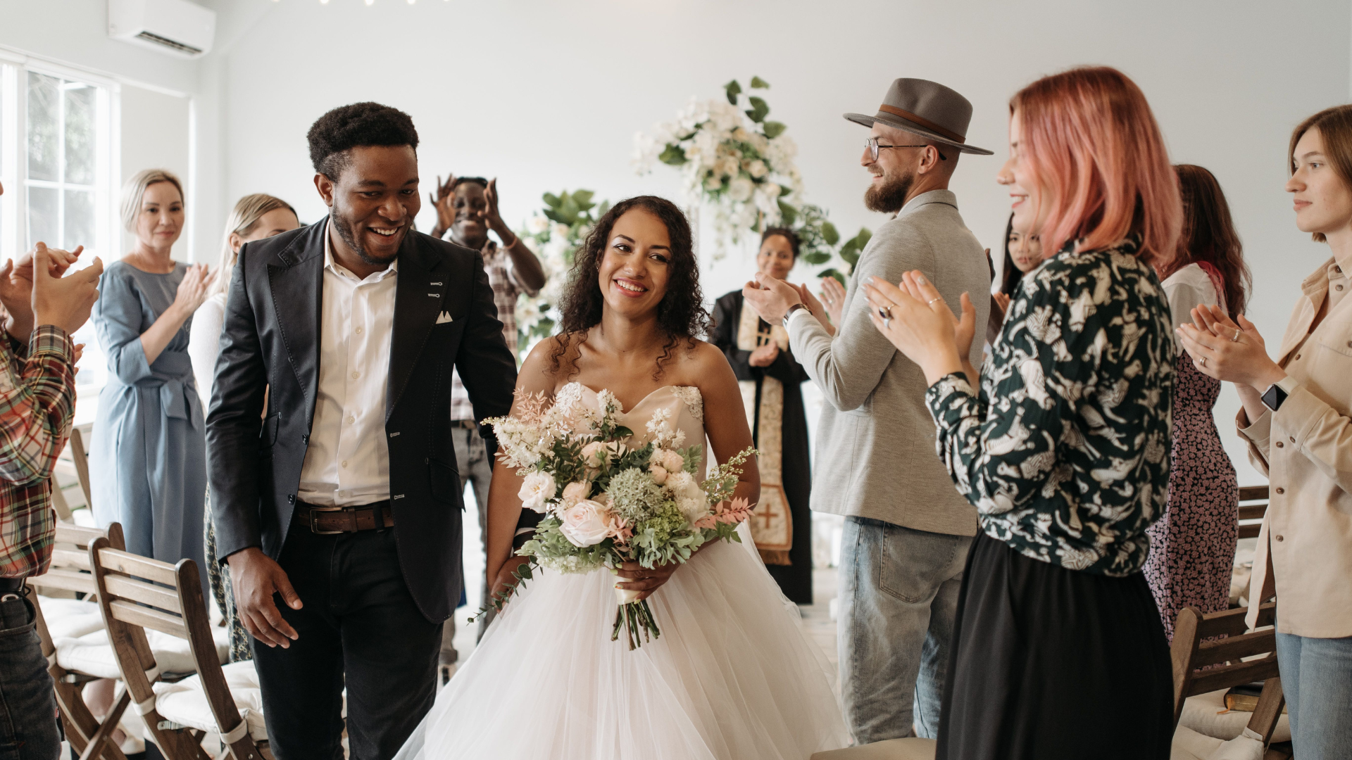 A smiling bride and groom walk down the aisle as wedding guests stand and applaud them in a brightly lit room.