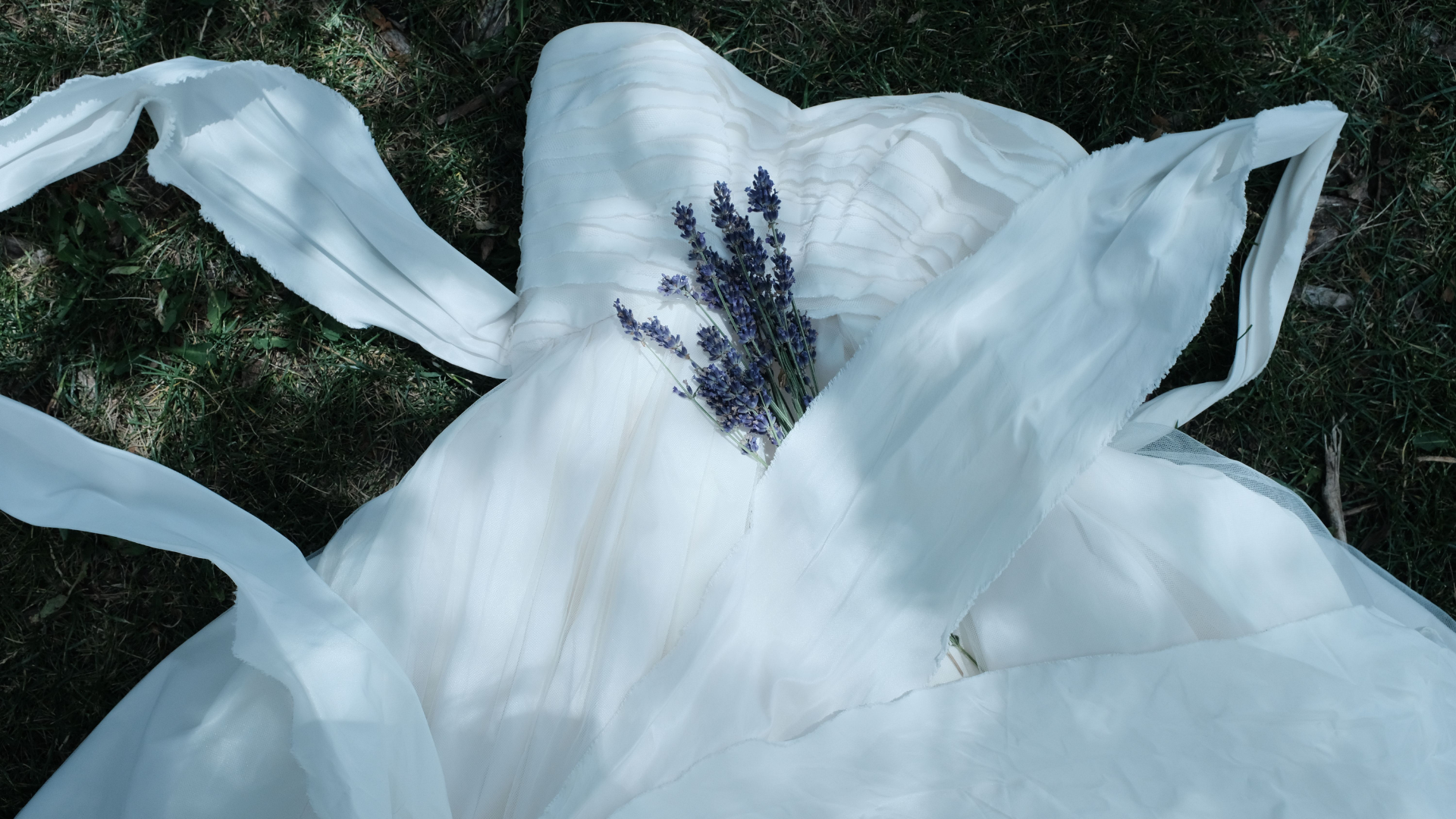 A white wedding dress is laid out on green grass with a small bouquet of lavender resting on the bodice.