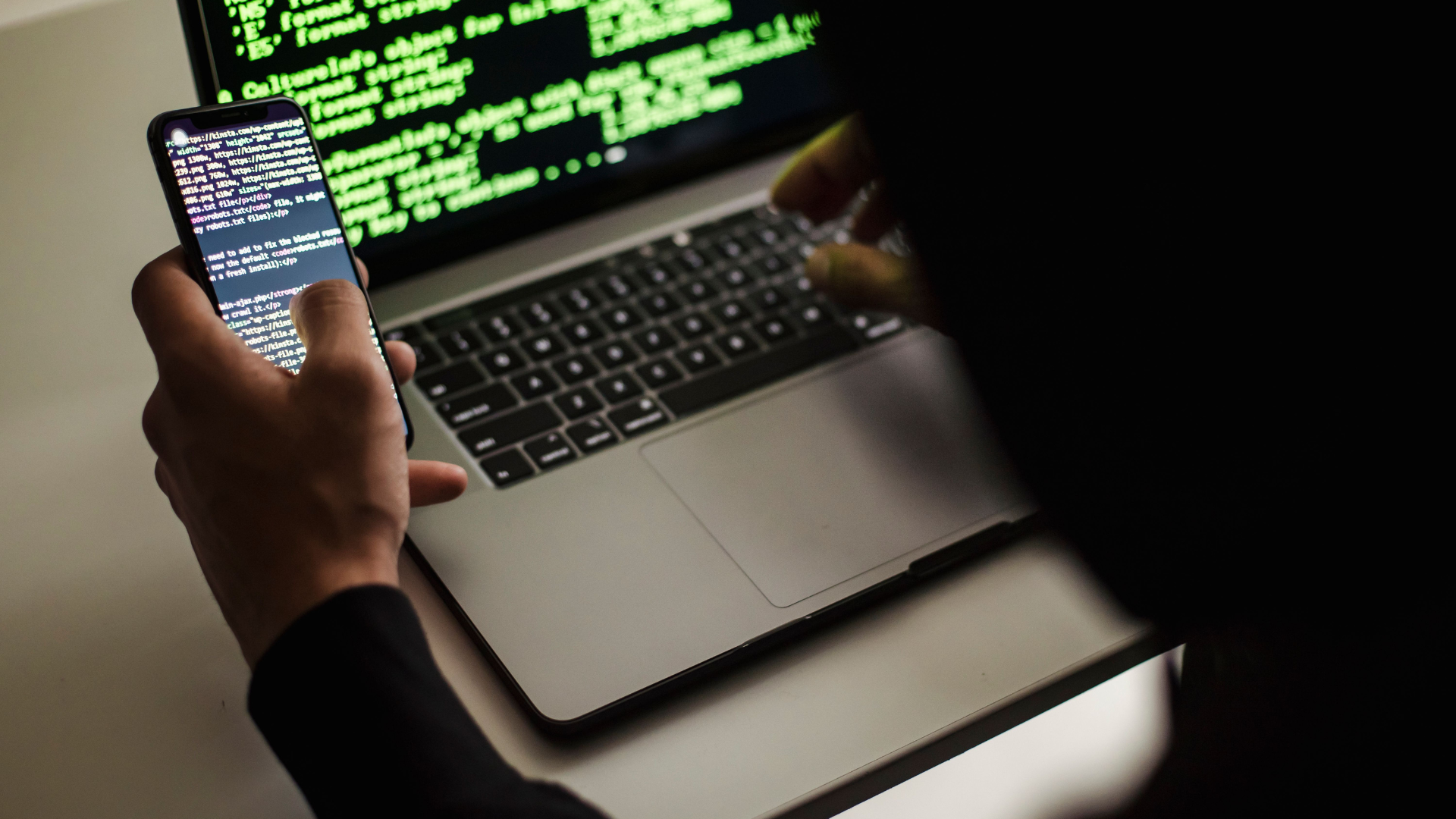 An over-the-shoulder view of a person using a smartphone and a laptop, both displaying lines of green computer code on black backgrounds.