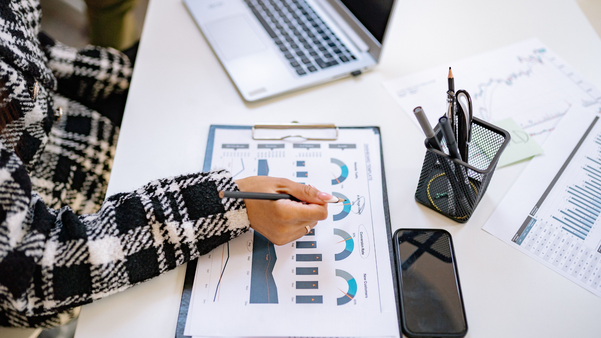 An overhead view of a person wearing a black and white plaid jacket holding a pencil over data charts on a clipboard alongside a laptop, smartphone, and desk organizer.
