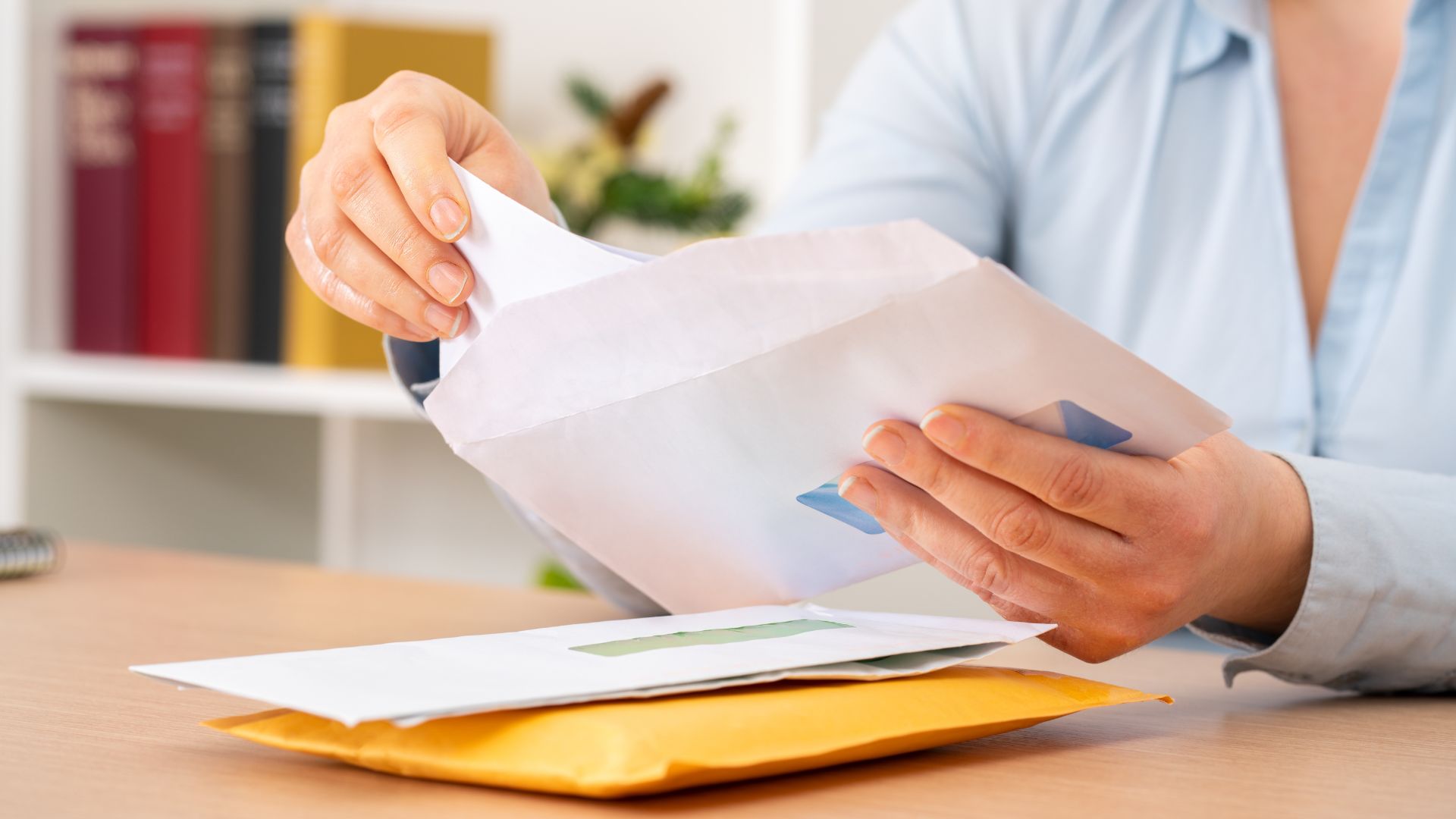 A person opening mail envelope at a wooden office desk with bookshelf behind.