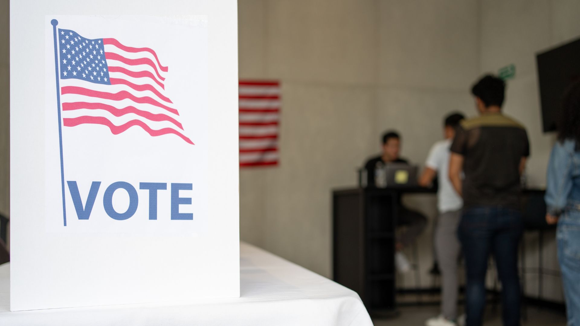 Polling station vote sign with American flag and voters in background.