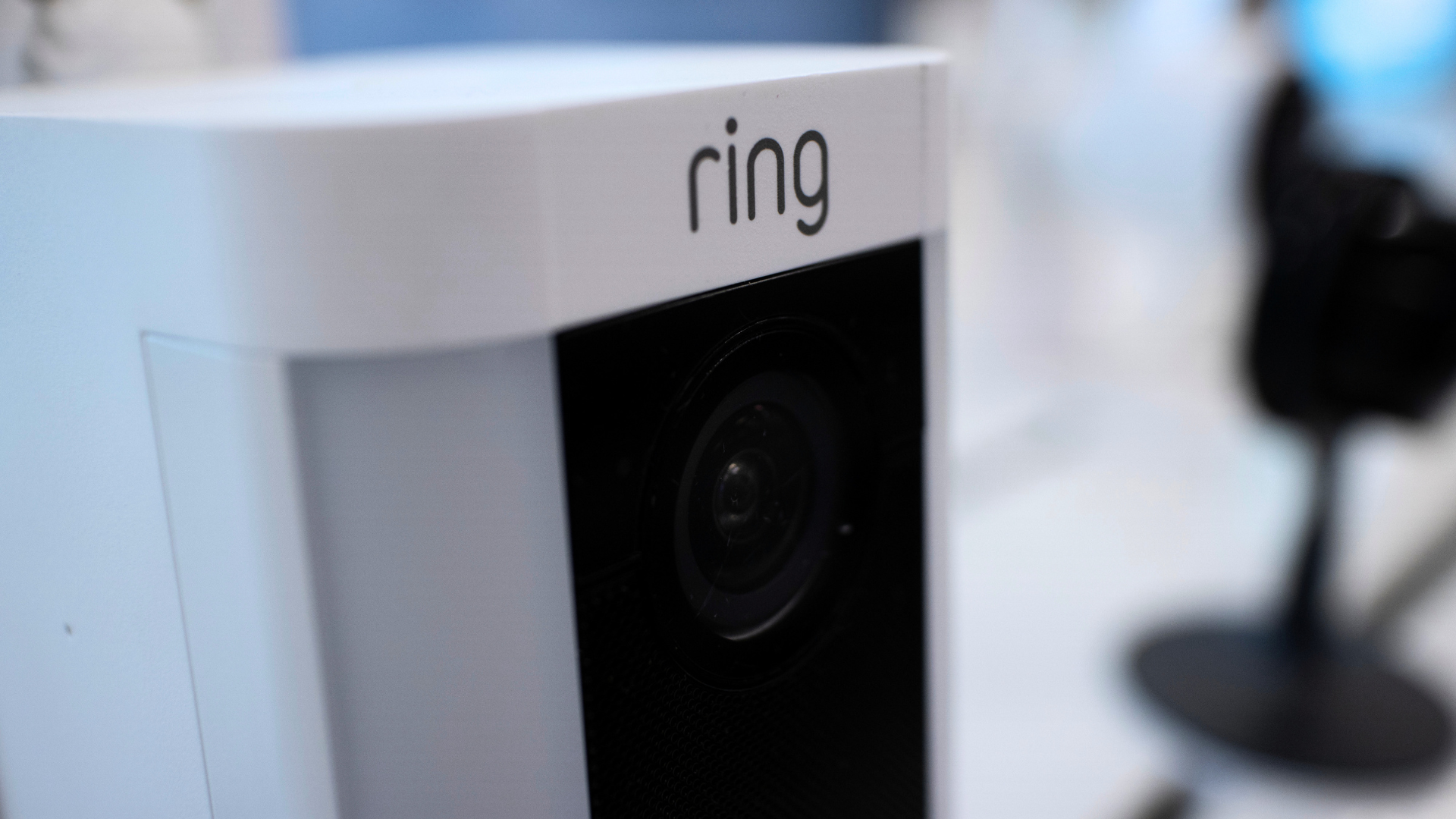 A close-up shot of a white Ring smart security camera, highlighting the dark lens housing and the prominent grey logo on top.