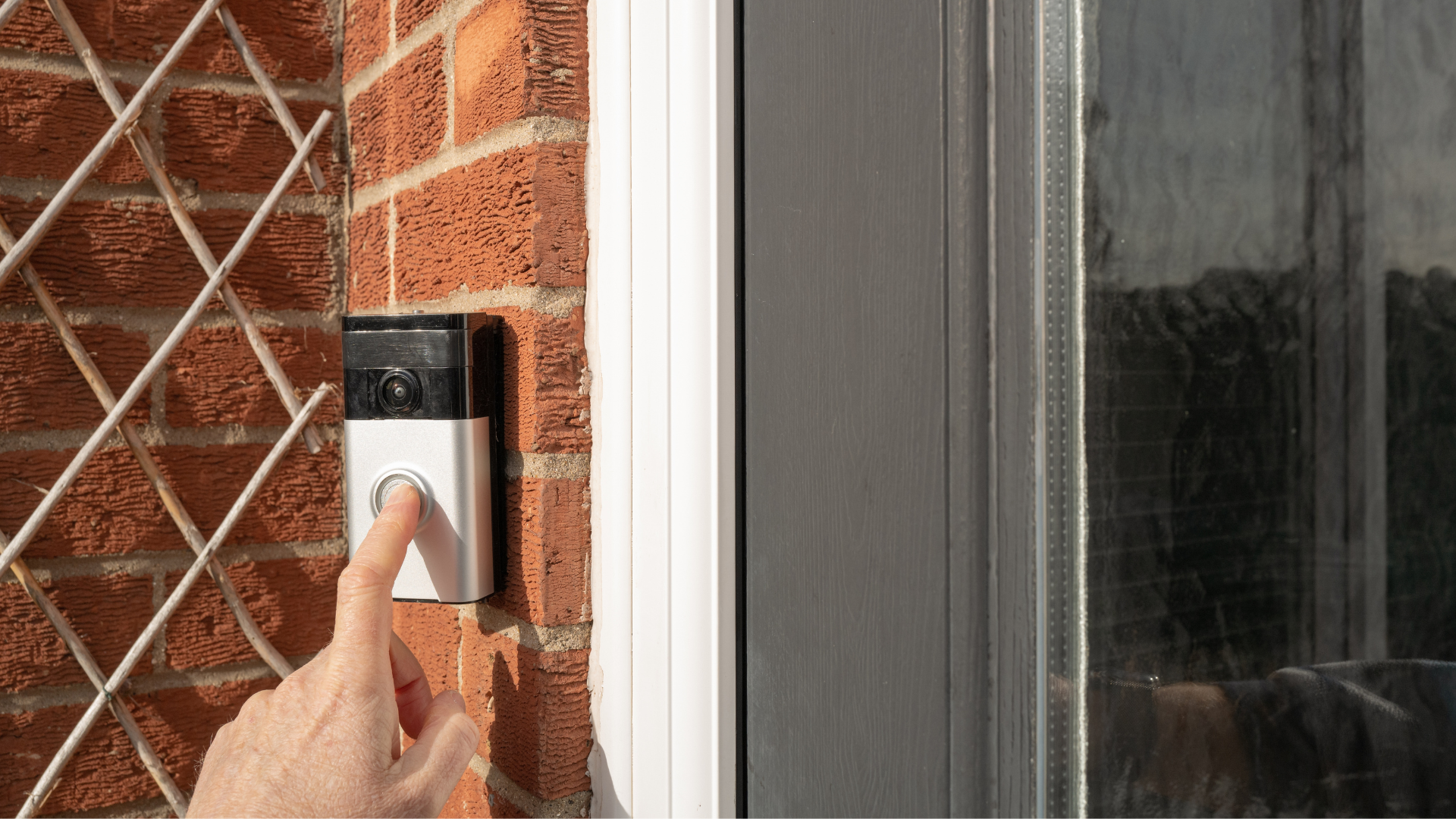 A side angle of a person's finger pressing the button of a silver-and-black smart video doorbell mounted to an exterior red brick wall.