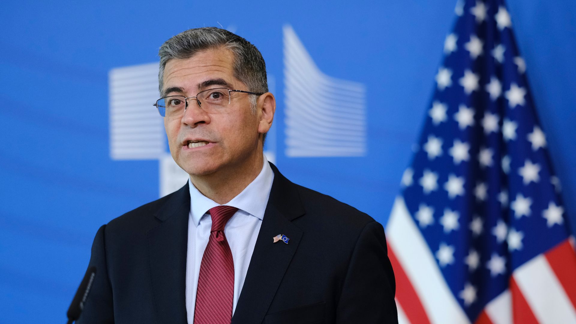 Xavier Becerra speaking at a press conference with the American flag in background.