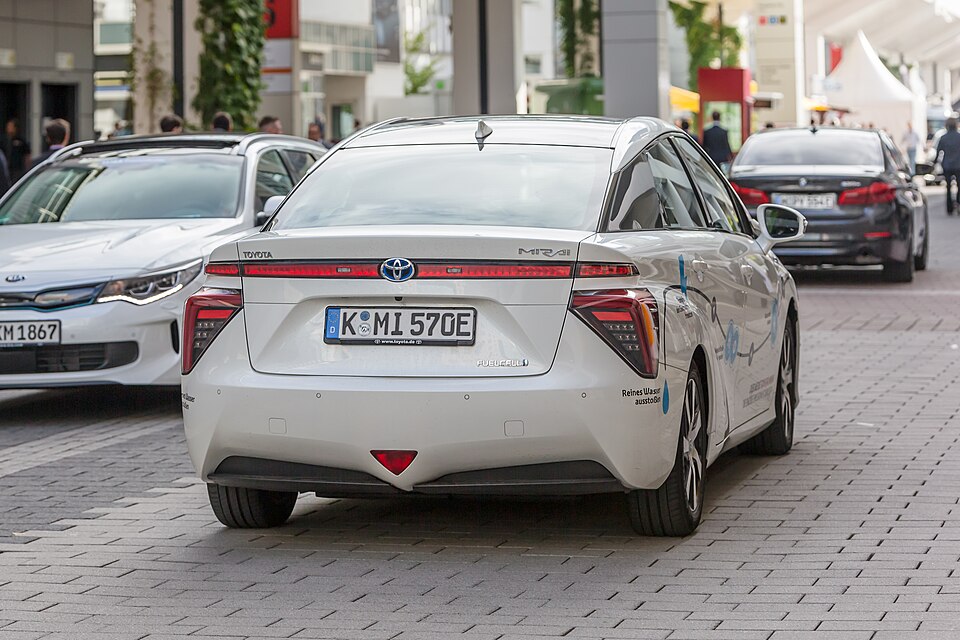 Toyota Mirai hydrogen fuel-cell vehicle driving in an urban area, representing the automaker's long-term investment in hydrogen-powered transportation.